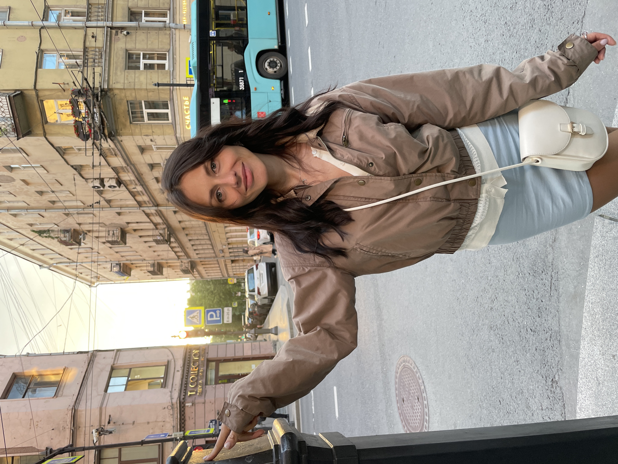 A woman is standing on a city street in Saint Petersburg, Russia. She has long, dark hair and is wearing a brown jacket over a white shirt and a light blue skirt. She is carrying a small white purse. The woman is standing next to a black pole, and her left hand is resting on it. In the background, there are buildings, a blue bus, and cars on the street. The sky is visible with sunlight. The scene appears to be in the evening.