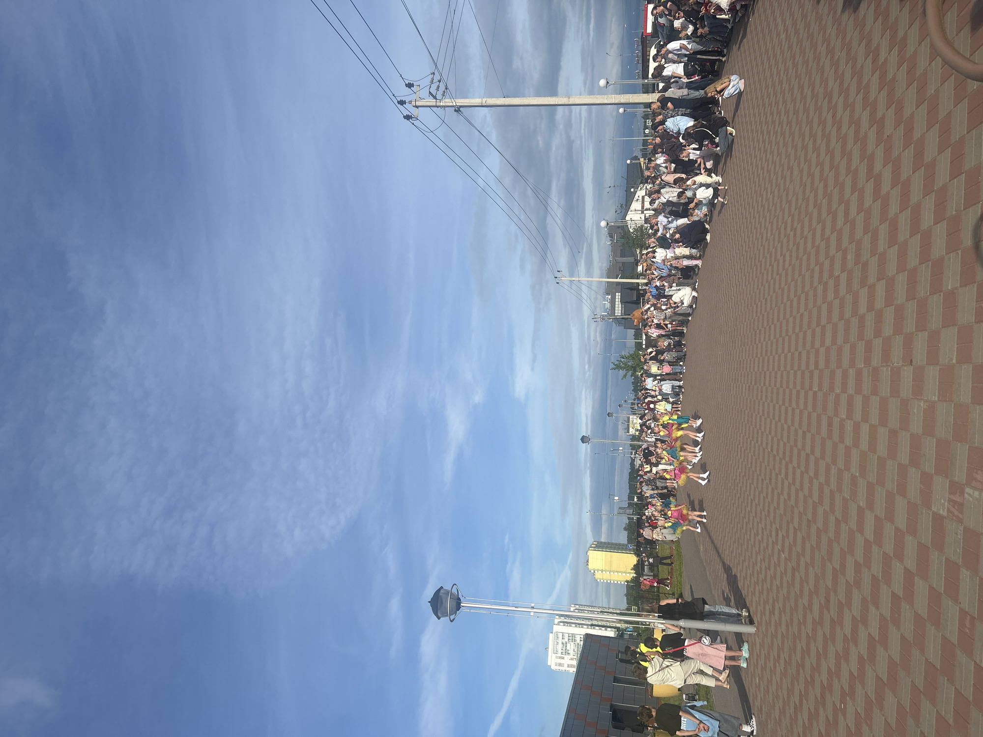 In a public outdoor area with a checkered brick walkway, a crowd of people is gathered under a partly cloudy blue sky. A group of young girls in colorful dresses, possibly performers, are in the center of the walkway, facing a larger group of spectators. To the left, a modern building with orange and gray panels is visible, along with a tall lamppost. In the background, several residential buildings and utility poles with power lines can be seen. The scene appears to be a community event or performance.