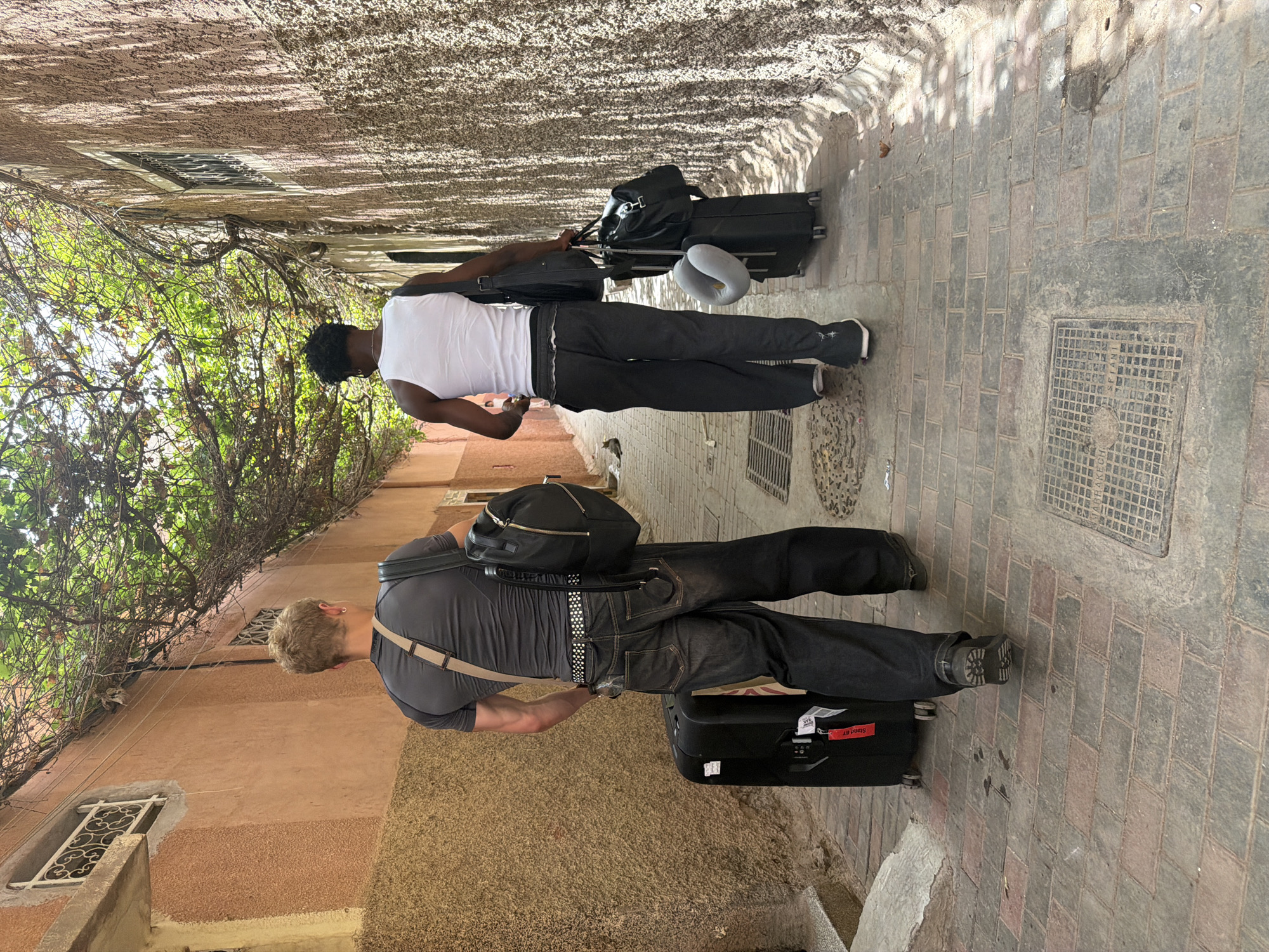 In Marrakesh, Morocco, two men walk away from the camera down a narrow alleyway. The alley walls are rough-textured and beige, with some green vines overhead creating dappled shade. The ground is paved with rectangular bricks. The man on the left, with blonde hair, wears a dark grey t-shirt, dark jeans, and a black backpack. He is pulling a black suitcase. The man on the right, with dark skin and hair, wears a white tank top, dark grey pants, and carries a dark bag over his shoulder and a black duffel bag on top of a rolling suitcase. A grey neck pillow is attached to the suitcase.