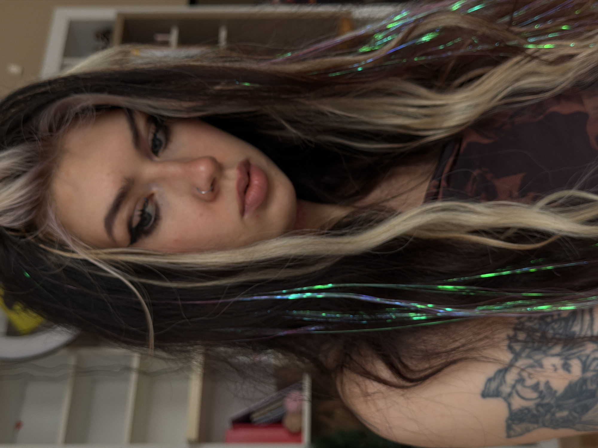 A medium close-up shot features a young woman with a fair complexion, dark eyeliner, and a nose ring. She is positioned at an angle, with her gaze directed towards the camera. Her hair is dark with blonde streaks and iridescent green and blue strands woven in. A section of her right shoulder is visible, displaying a tattoo. She appears to be wearing a dark brown patterned garment. The background shows a blurry view of what appears to be a room with white shelving units. The image provides no indication of being in Budva, Montenegro, or any specific city.