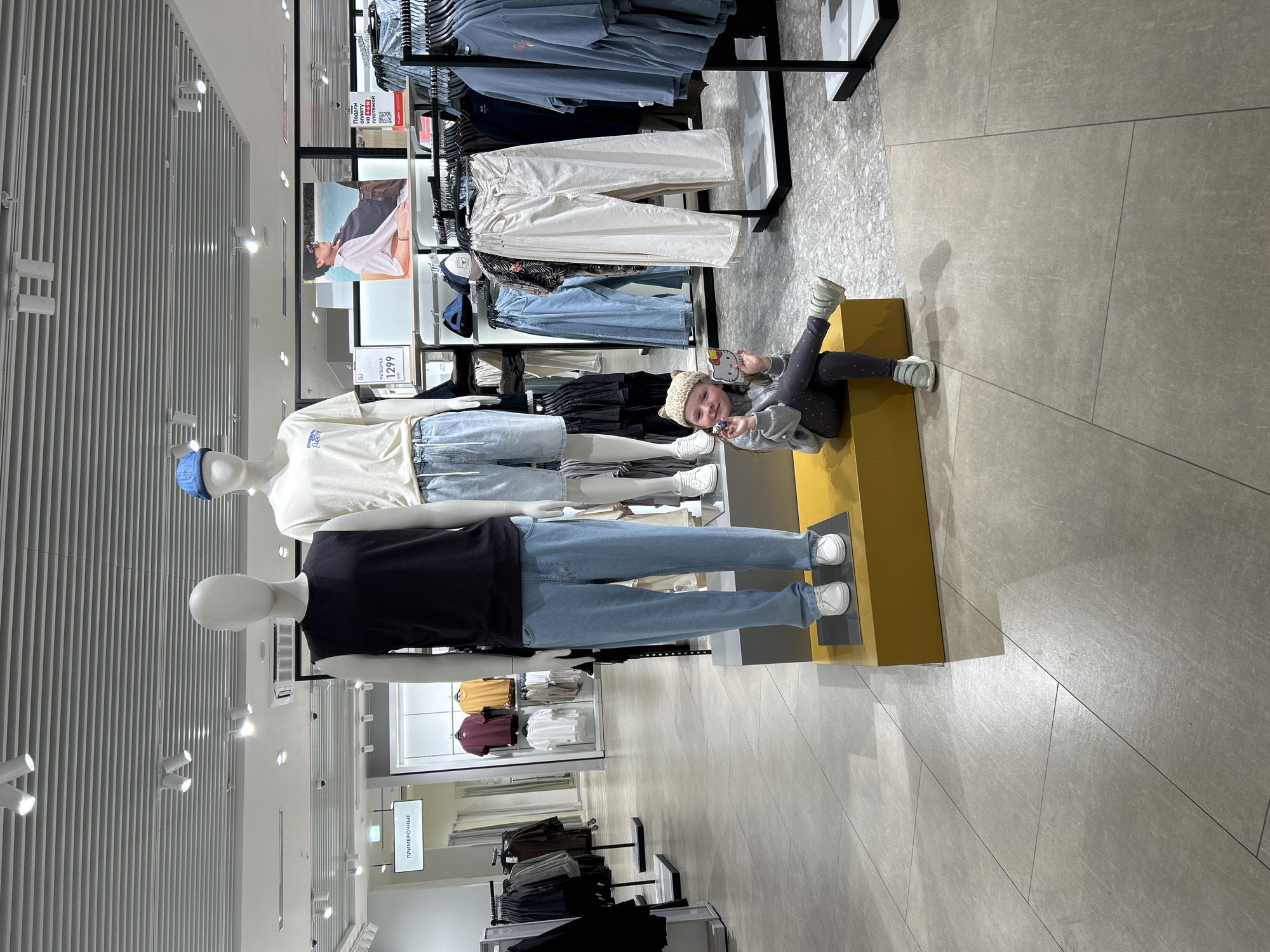 The image captures the interior of a clothing retail store located in Yekaterinburg, Russia. The store features a light gray tiled floor with visible grout lines, transitioning to a different gray textured or terrazzo-like flooring further into the store on the right. The ceiling is white and slatted, equipped with numerous track lights, recessed lights, and several black dome-shaped security cameras. Walls are light-colored, likely white or light gray.
In the central foreground, three headless mannequins are displayed on two interconnected yellow rectangular platforms. The leftmost mannequin wears a black sleeveless top, light blue jeans, and white sneakers. The middle mannequin wears a cream-colored short-sleeve t-shirt with blue text, light blue denim shorts, and white sneakers. A third mannequin, partially visible on the right, wears light blue shorts and white sneakers.
Behind the mannequins, black metal clothing racks are filled with various garments, including t-shirts, pants, and shorts in shades of black, white, cream, blue, burgundy, and yellow. Shelves below a large wall poster also display folded clothing and several hats.
Signage visible in the store includes a hanging sign with white text on a black background reading "ПРИМЕРОЧНЫЕ" (Fitting Rooms). Another sign indicates a price, "ФУТБОЛКА 1299 руб." (T-shirt 1299 rubles). A promotional sign, "Подели оплату на 4-10 платежей" (Divide payment into 4-10 payments), features a QR code. A large wall-mounted poster depicts a man wearing a white and black collared shirt.
A young female child is seated on the front yellow display platform, to the right of the central mannequin. She has light-colored hair and is wearing a patterned beige and brown beanie, a light gray hooded sweatshirt, dark gray leggings with light polka dots, and light green sneakers. Her left leg is bent with her foot on the platform, and her right leg is extended forward with her foot resting on the floor. She is looking towards the camera with a slight smile. In her left hand, she holds a small, dark-colored, possibly metallic figurine. In her right hand, she holds a white, heart-shaped object featuring a pink Hello Kitty design.