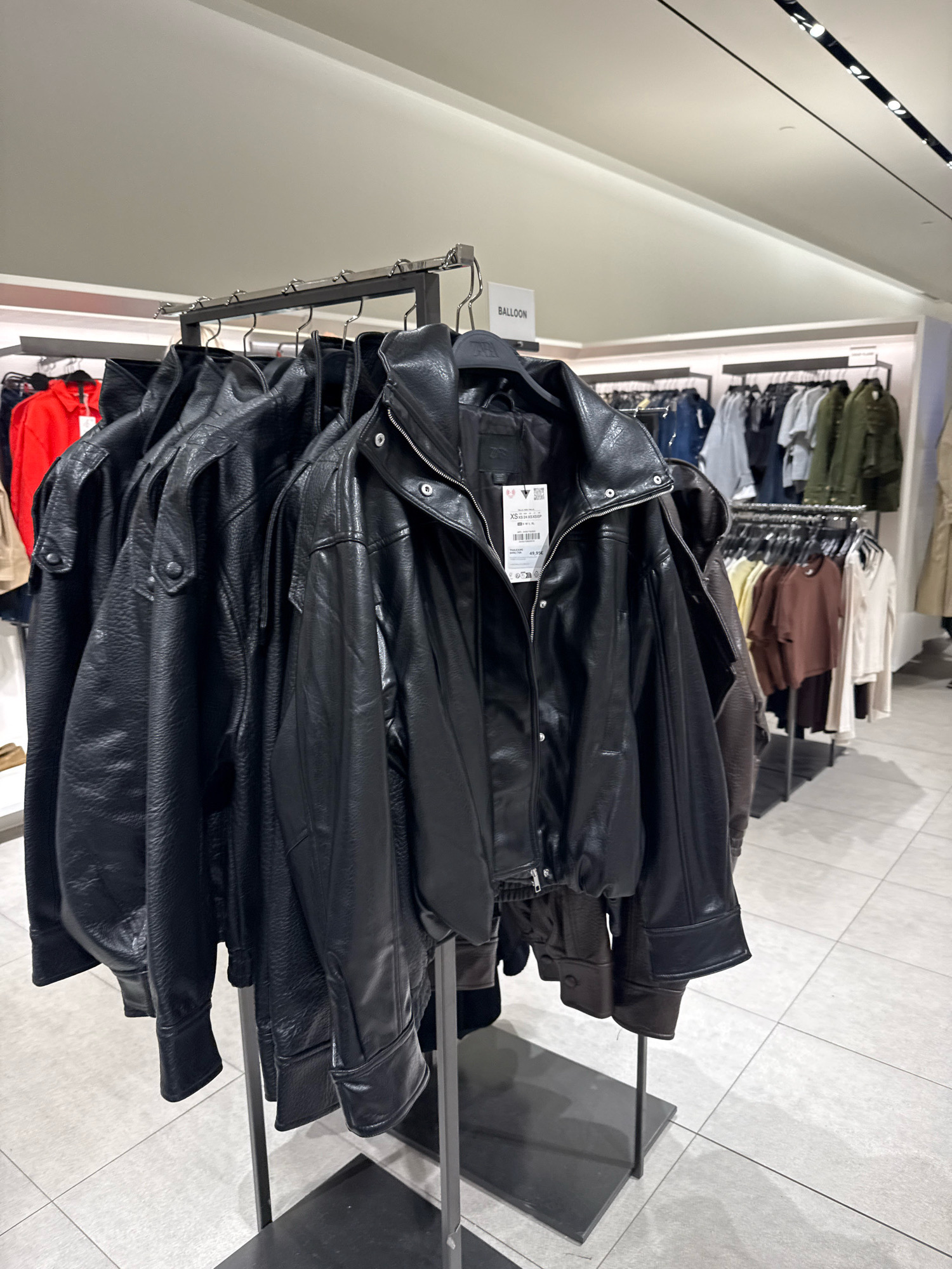 In a retail store, several black faux leather jackets hang on display racks. One jacket in the foreground has a white tag with text and a barcode. Behind the jackets, other clothing items are visible on racks in the background, including red, blue, white, brown, and green garments. The store has a tiled floor and overhead lighting. The context indicates this scene is in a clothing store in Tallinn, Estonia.