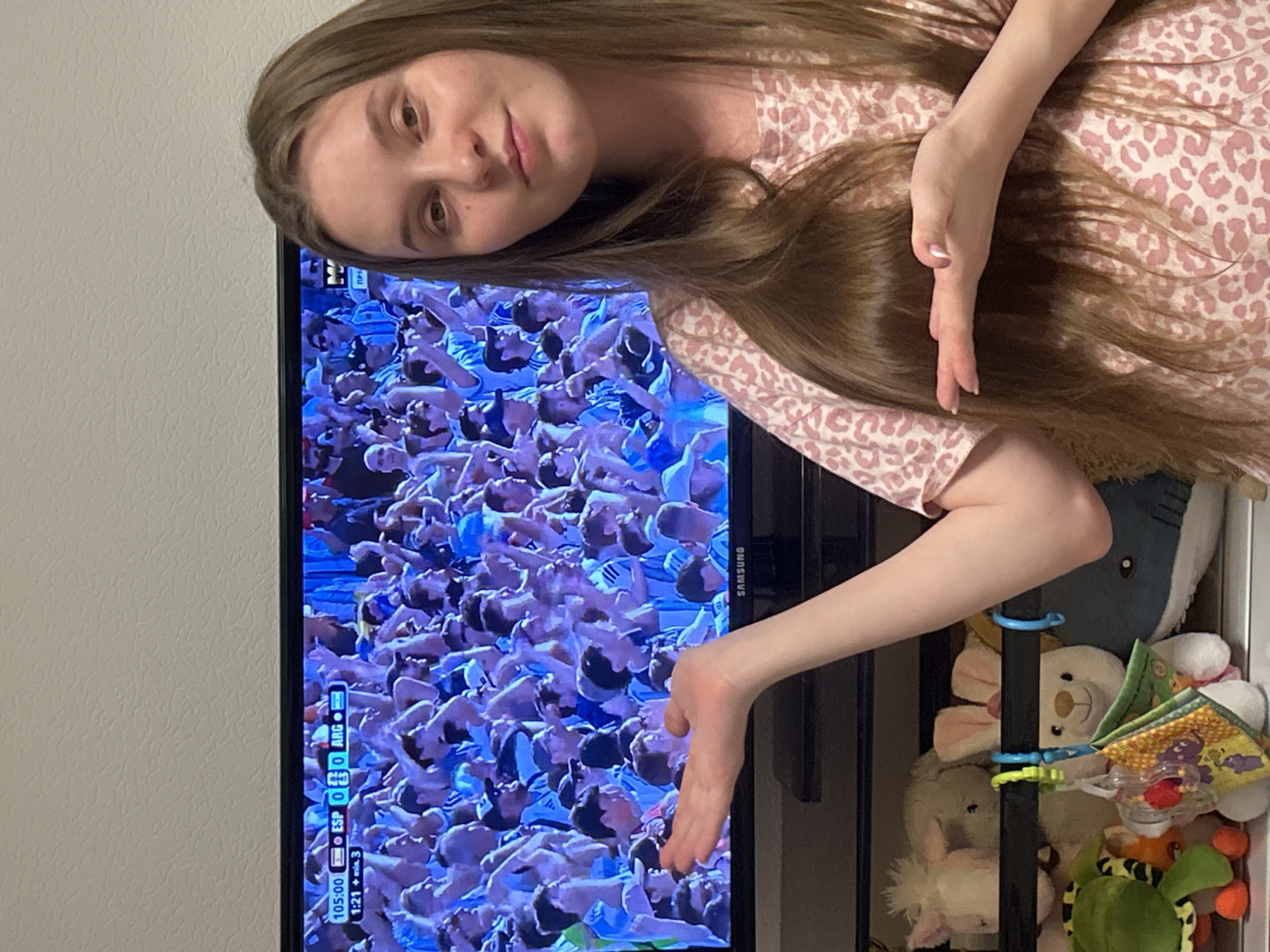 A young woman with long, light brown hair stands in front of a television screen displaying a soccer match. She is wearing a pink top with a leopard print pattern. Her left hand is extended and open, palm facing upward, towards the television, while her right hand is also extended, palm facing upward. The television screen shows a crowd of people in a stadium wearing blue and white. A scoreboard at the top of the screen indicates "105:00 ESP 0 ARG 0". Below the screen, a shelf contains several stuffed animals. The background wall is textured and painted a light color. The image appears to be taken indoors, likely in a home setting in the city of Noviy, Russia.