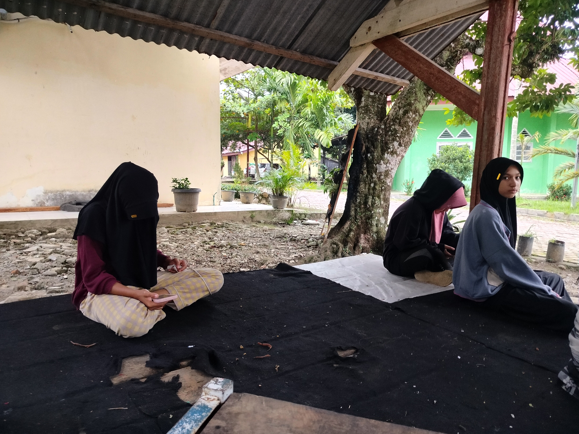 In Purwodadi, Indonesia, three young individuals are seated outdoors on a black mat beneath a corrugated metal roof supported by wooden beams. The scene appears to be an open-air shelter with a light-colored wall on the left and lush green foliage and trees visible in the background, suggesting a tropical climate.
On the left, a person wearing a black niqab and a burgundy sweater with checkered pants is seated cross-legged, holding a pink object, possibly a phone, in their hands. To their right, another individual in a black hoodie with a pink headscarf and dark pants is also seated. Further to the right, a third person, wearing a black hijab and a light blue hoodie over black pants, is looking towards the camera. The ground is covered with debris and gravel. In the background, there are potted plants, a large tree with mossy bark, and buildings with green and yellow walls. The overall atmosphere suggests a casual gathering or study session in an outdoor setting.