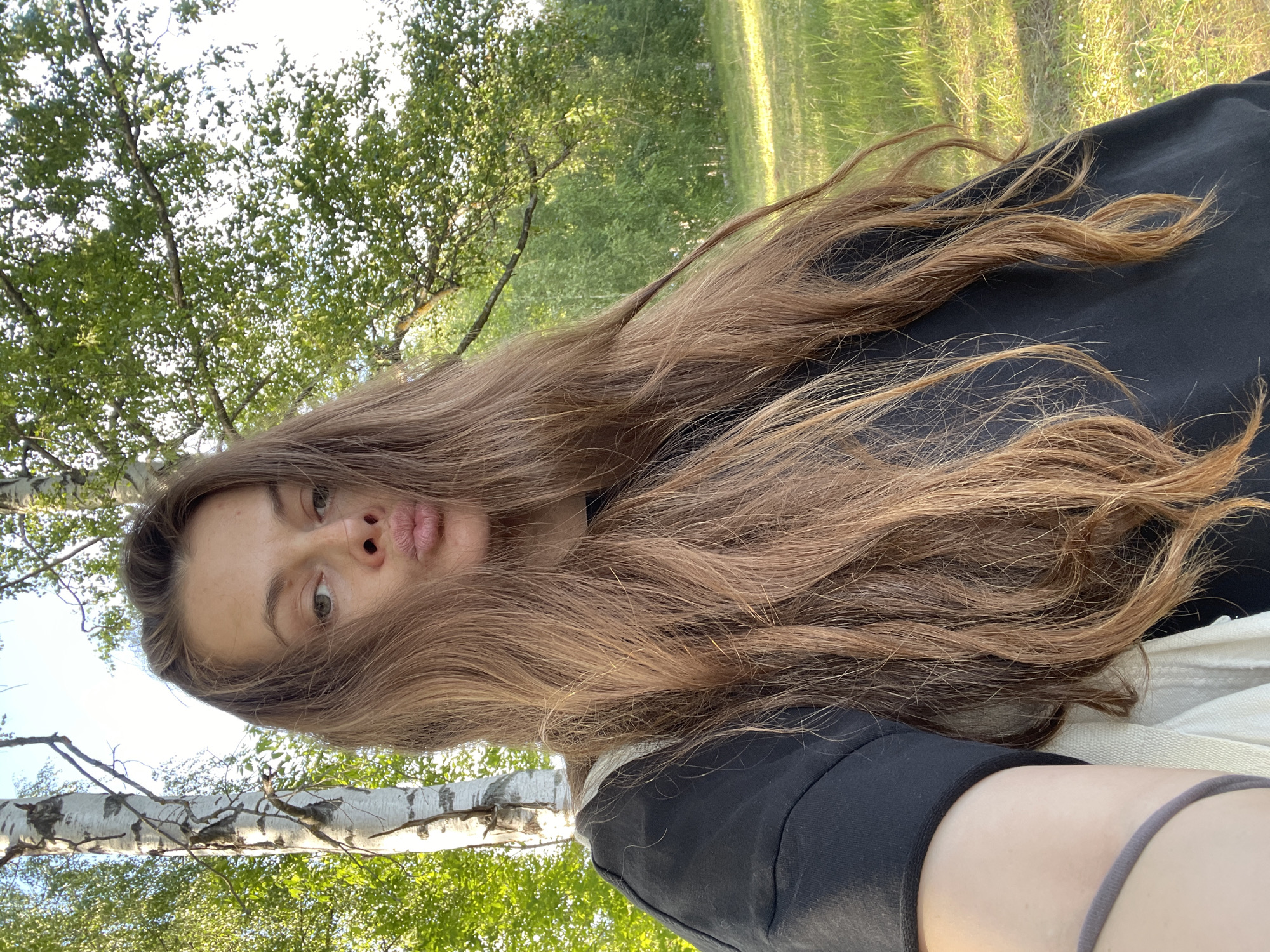 A young woman with long, light brown hair is captured in a low-angle, outdoor selfie. Her hair is draped across her face and shoulders, obscuring much of her body. She is wearing a black top with a white strap visible. The woman is making a kissing face towards the camera.
The background suggests a park or a wooded area with green grass and trees. Sunlight filters through the leaves, creating dappled light on the ground. There are birch trees with white, mottled bark visible in the foreground and background. The time of day appears to be late afternoon or early evening, given the warm, golden light. The weather seems pleasant and sunny. The location context provided is Заречный, Russia. No visible text is present in the image.