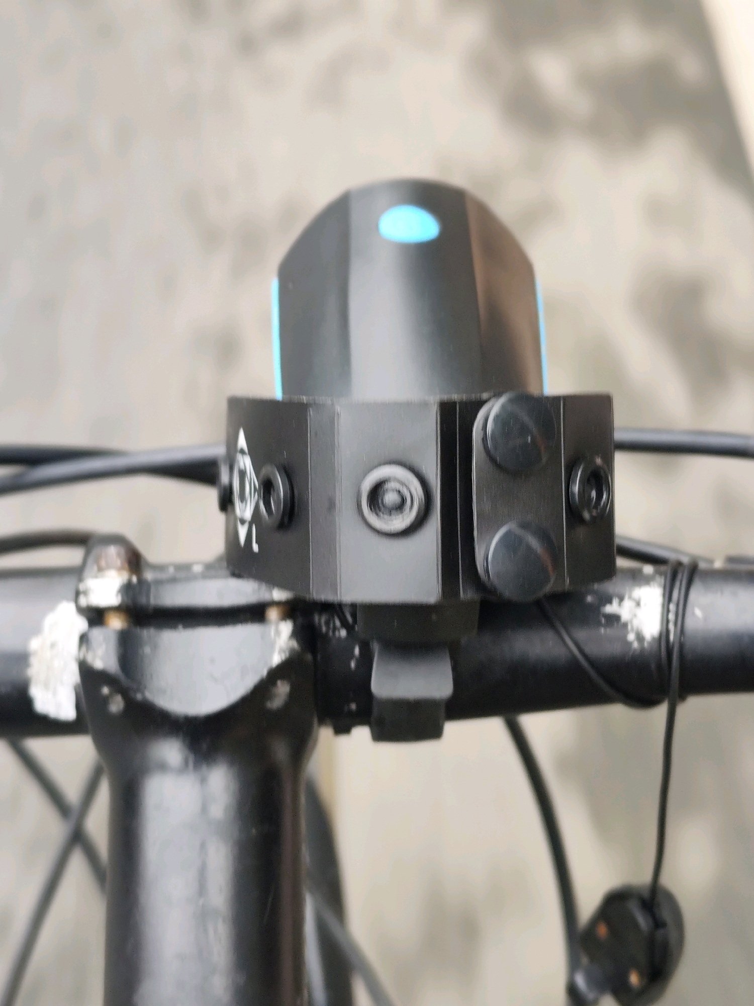 A close-up, slightly blurry, eye-level shot shows a black bicycle handlebar with a black and blue electronic device clamped to it. The device appears to be a bicycle light or bell. The handlebars are black metal, and the device has several black screws visible. To the left of the device, a white triangular symbol with a diagonal line is visible. The background is out of focus and appears to be a light-colored, textured surface, possibly a wall or pavement. The image does not provide any specific landmarks or context to indicate it is located in Da Lat, Vietnam.