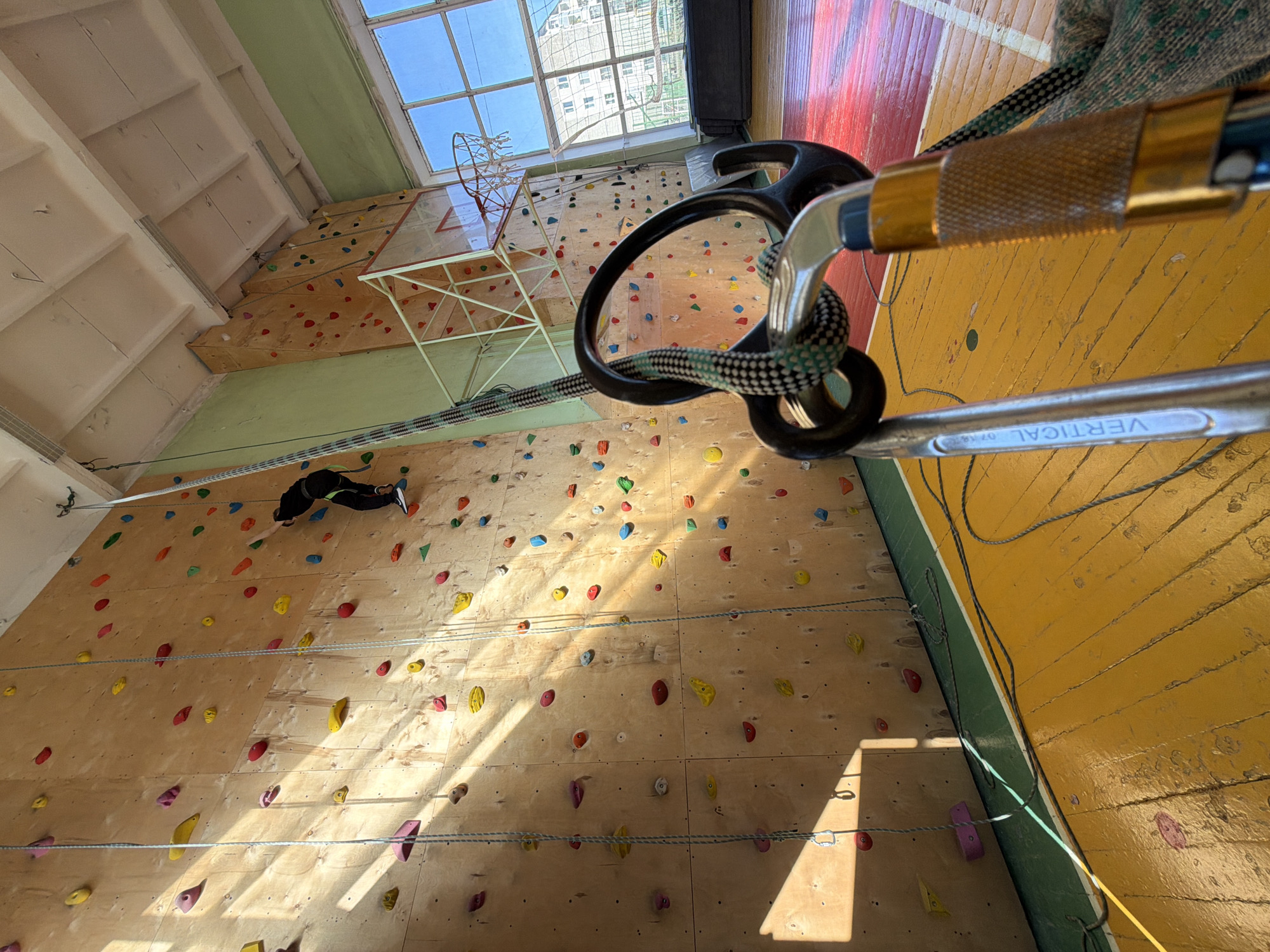 A high-angle shot depicts a climbing wall at an indoor facility, with a climber visible near the top left. The climbing wall is wooden, with a variety of colorful climbing holds attached. A rope and belay device are in the foreground, suggesting the scene is focused on climbing safety and equipment. A basketball hoop is visible in the background, suggesting this may be a gymnasium-style space. The location is Izhevsk, Russia.