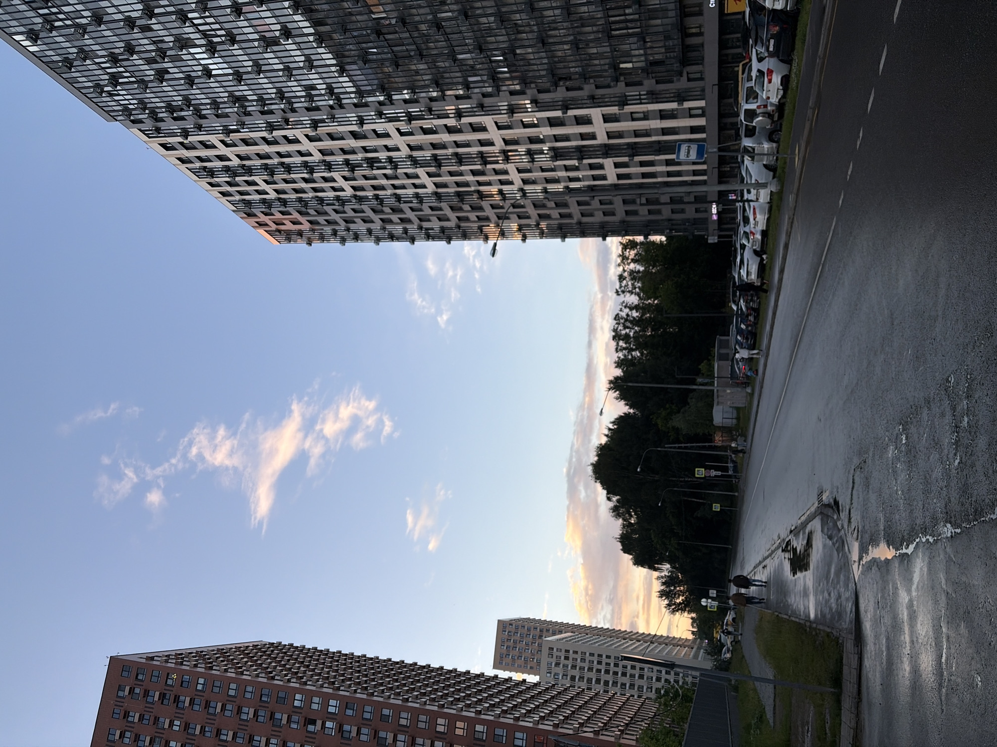 The image depicts a street scene in the city of Khimki, Russia, during twilight. Two tall apartment buildings dominate the skyline, one on the left constructed of red brick and another on the right clad in glass and dark metal. A smaller, lighter-colored apartment building is visible between them. A wet asphalt road runs diagonally across the frame, with a sidewalk on the left. Several vehicles are parked along the right side of the road. Two individuals are walking on the sidewalk, appearing as silhouettes against the backdrop. Puddles of water are visible on the wet road and sidewalk, reflecting the soft light of the sky. Trees line the far side of the road, and a streetlamp is visible near the right building. The sky is a gradient of blue and orange, with wispy clouds tinged by the setting sun.