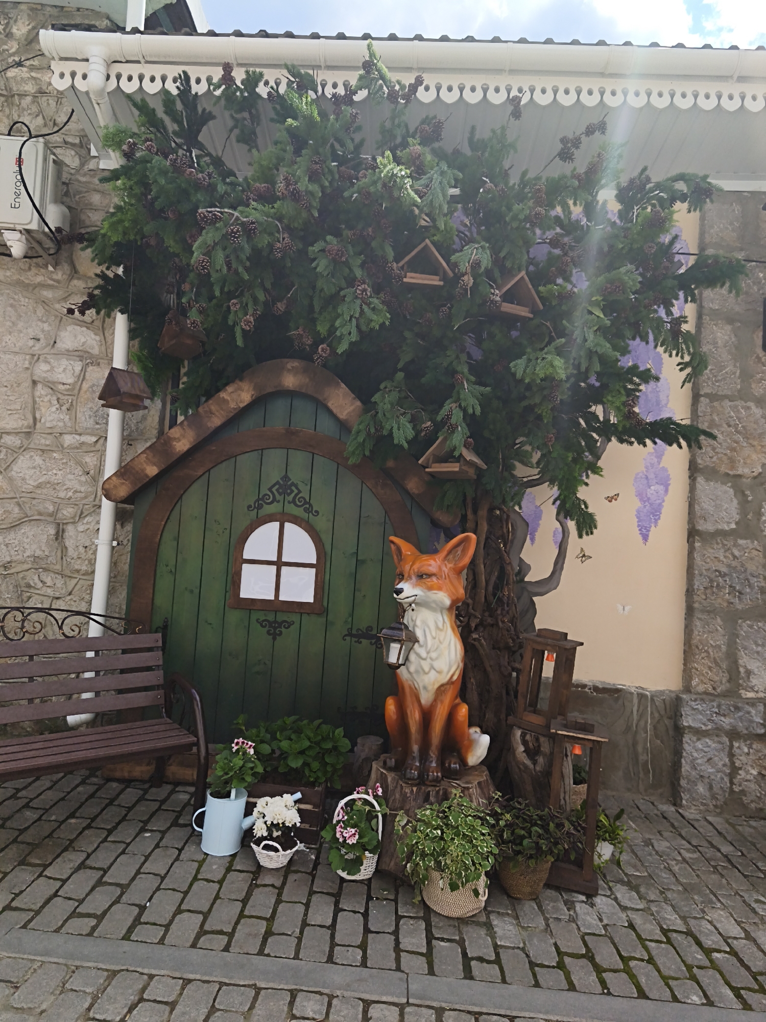 The image depicts a whimsical outdoor scene, likely in the city of Алупкинский городской совет, Ukraine, given the location context. At the center is a faux hobbit-style house with a green arched door and a small, multi-paned window. Overhanging the house is a dense evergreen tree adorned with pine cones and birdhouses. To the left, a simple wooden bench sits on a stone-paved ground. A decorative fox statue holding a lantern is positioned to the right of the hobbit-house door, surrounded by various potted plants and small floral arrangements. To the right of the statue, wooden frames are stacked against a stone wall, which transitions into a mural depicting a woman reaching for wisteria flowers and butterflies. The overall scene suggests a curated, storybook-like aesthetic in an urban environment.
