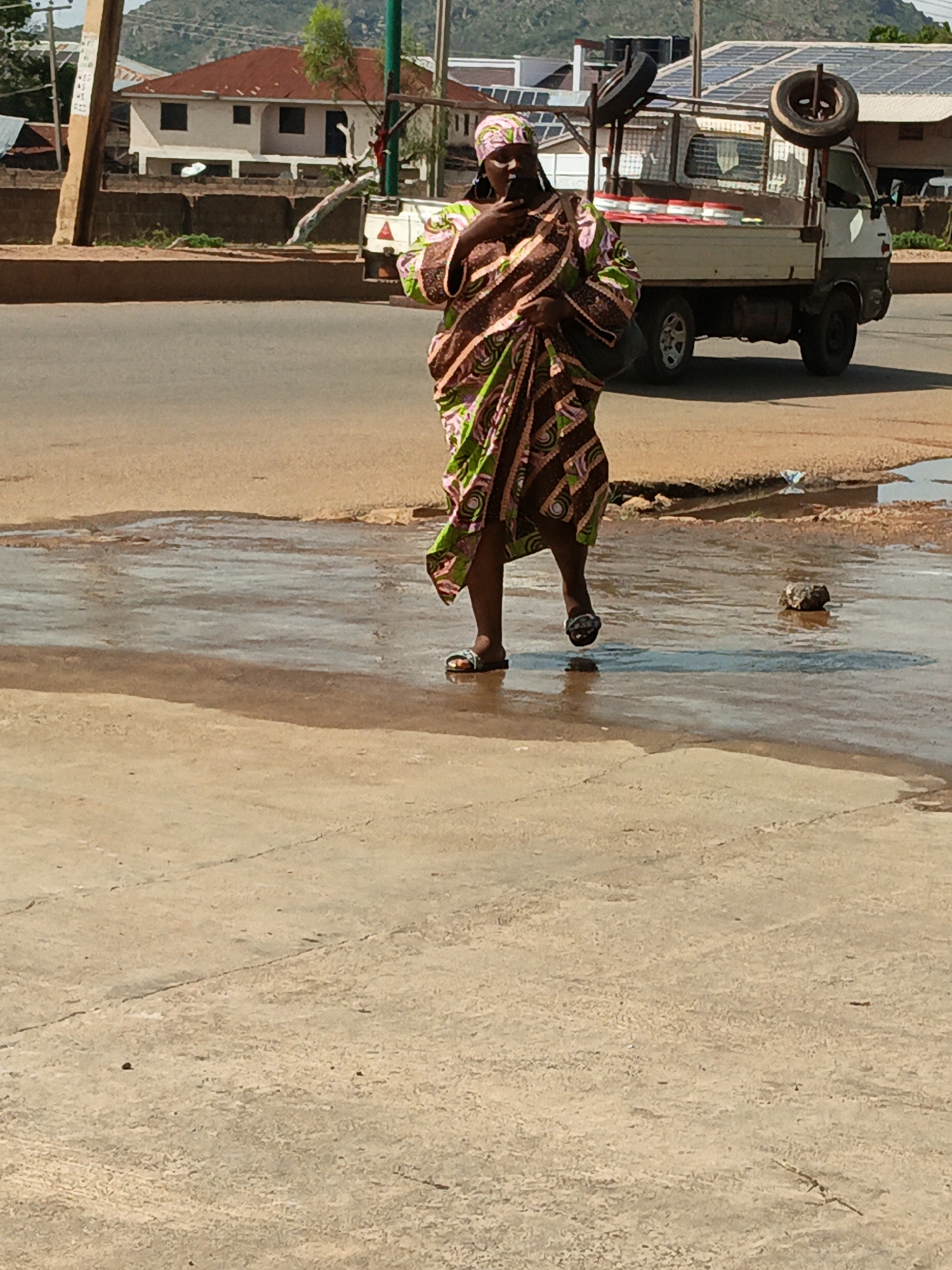A woman in a patterned brown and green dress and headscarf walks across a wet, paved street, carrying a dark bag on her shoulder. She wears sandals. A white truck with a cage-like structure and two tires mounted on it is parked in the background. Buildings, trees, and a mountainous landscape are visible further behind. The scene appears to be an outdoor street environment with signs of recent rain.