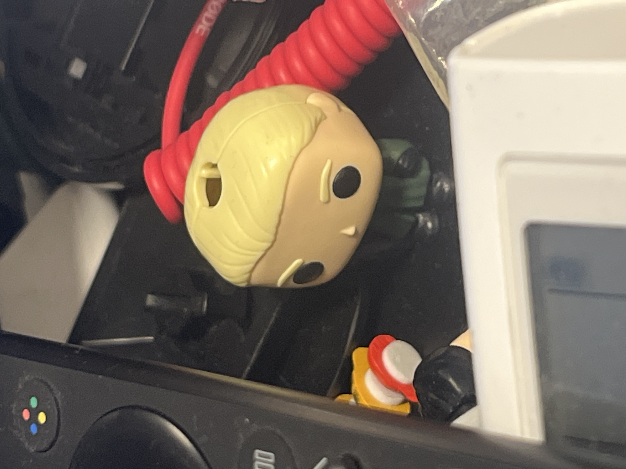 This close-up image shows a collection of electronic accessories and a collectible figure, likely situated on a desk or shelf in Tel-Aviv, Israel. The central subject is a blonde-haired Funko Pop figure, light-skinned with large black eyes, lying on its side. A bright red coiled cable, bearing the partially visible text "RODE" (suggesting an audio equipment brand), curves around the top of the figure's head.
In the lower left corner, a dark grey/black video game controller is visible, featuring a distinct four-color button cluster (red, blue, green, yellow dots) and a large black circular button or analog stick, indicating a retro console or themed controller. Below the Funko Pop, to the right, a small item composed of stacked red, white, and yellow circular pieces is present. A portion of a white electronic device or stand is visible on the far right, with clear plastic material in the upper right quadrant. A dark green object is partially seen behind the Funko Pop figure.
The scene is illuminated by artificial indoor lighting. No specific activity is occurring; the objects are static. The controller shows some signs of dust.