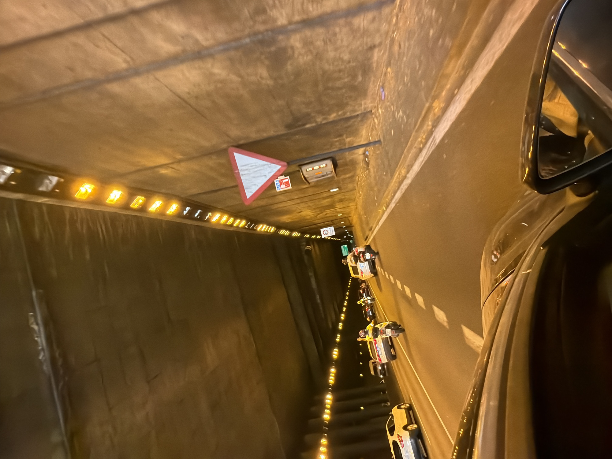 The image shows a view from inside a car, looking forward through the windshield and out the side mirror. The car is in traffic within a tunnel. The tunnel walls are visible and appear to be made of concrete. Along the top of the tunnel, there is a series of orange lights. The road surface is asphalt, with white dashed lines indicating lanes. Several other vehicles are visible on the road, including cars and motorcycles. One motorcycle rider is visible with their hand raised. There is a traffic sign on the right wall of the tunnel, a white triangle with a red border, indicating a yield or priority road. There is also a smaller sign below it with a red cross and an "SOS" symbol. The scene is illuminated by the tunnel lights and headlights of the vehicles, giving the image a warm, orange hue. The overall impression is a snapshot of everyday traffic within an urban tunnel environment, likely during the evening or night due to the artificial lighting. While the image itself doesn't explicitly state the location, the presence of a yield sign with a red border and triangle is common in many countries. Without further context or identifiable landmarks, it's impossible to definitively confirm the location as Barcelona, Spain, based solely on the visual information provided in the image.