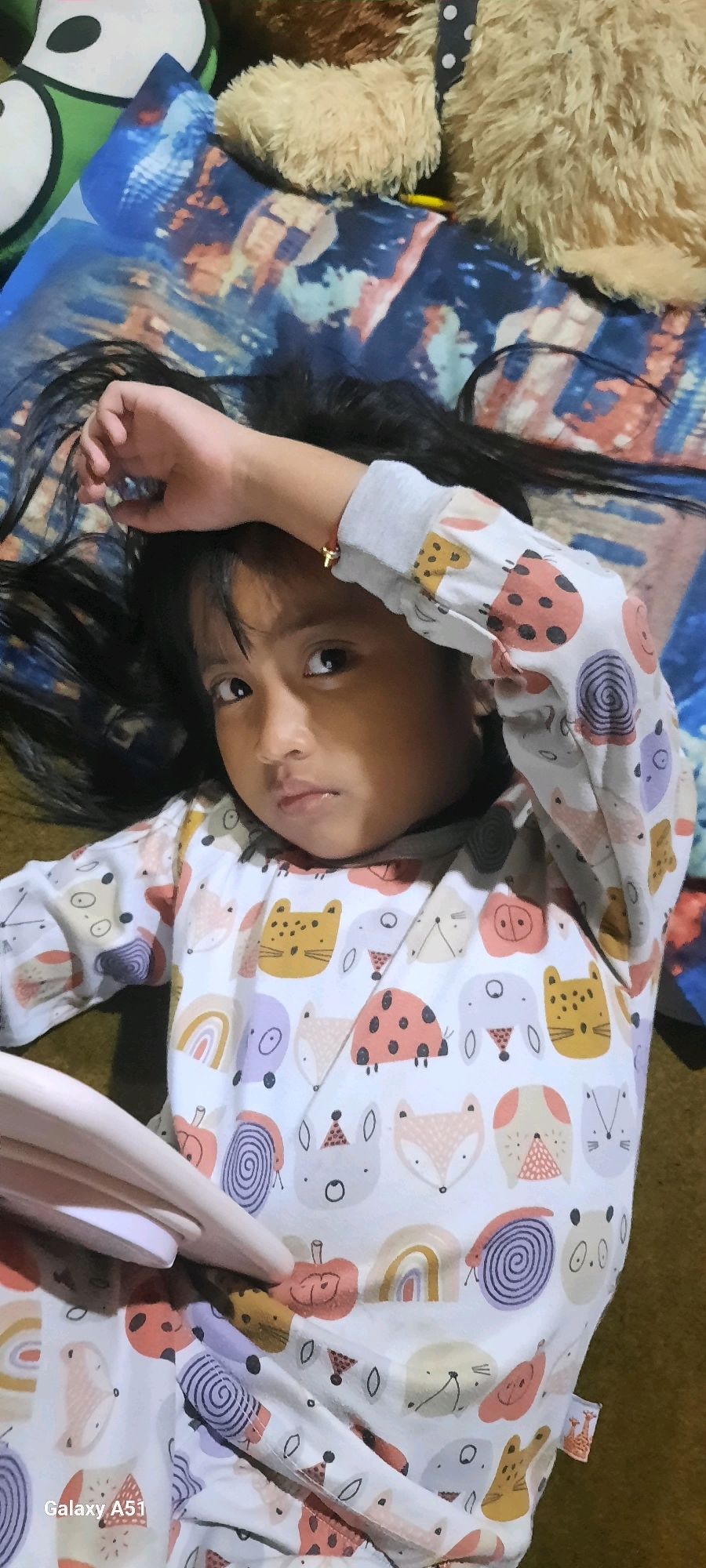 A young girl with long dark hair lies on her back on a patterned blanket, her right arm bent with her hand resting on her forehead. She wears a long-sleeved shirt with a whimsical print of cartoon animals and objects like snails, foxes, cats, and ladybugs. The blanket has a blue and orange abstract pattern. In the background, out of focus, are stuffed toys, one of which is fuzzy and tan. The setting appears to be indoors, possibly a bedroom. There is no visible indication of the specific location being Jatitengah, Indonesia, beyond the provided prompt.