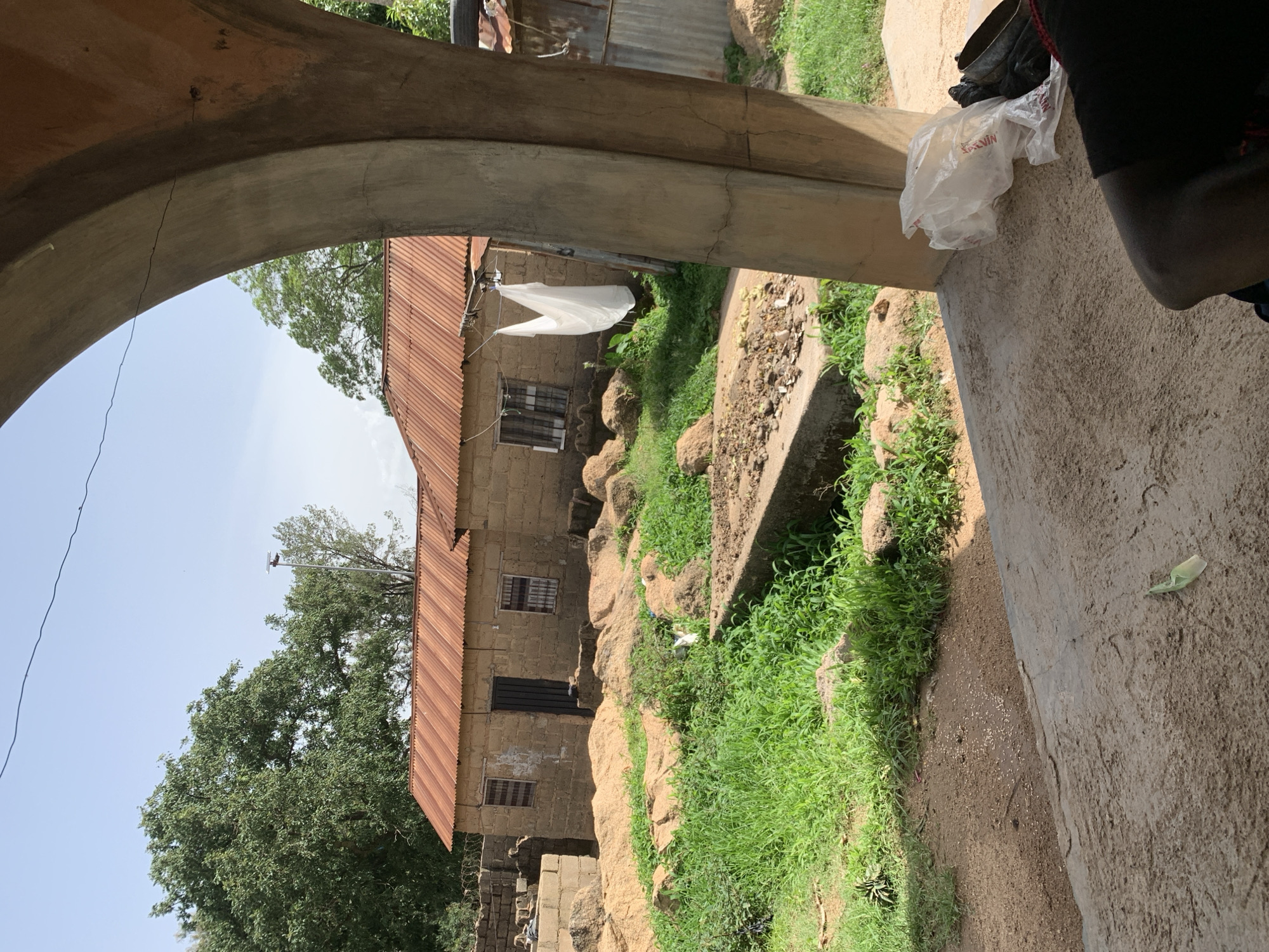 This is a medium shot of a rural scene in Jos, Nigeria, captured on a bright, overcast day. In the foreground, a thick, curved concrete pillar frames the right side of the image. A partially obscured person wearing a black shirt is visible in the lower right corner. Scattered on the concrete ground in front of the pillar are white plastic bags, one appearing to contain more bags, and a black bag. A second, flatter concrete structure runs horizontally across the mid-ground.
Beyond this structure, a modest, single-story building with rough, sandy-colored walls and a corrugated iron roof with a reddish-brown hue stands. Two windows with barred coverings and a dark door are visible on its front. A white piece of cloth is hanging from a line outside the building.
The area surrounding the building is rugged and uneven, with patches of green grass and scattered rocks. The ground is a mix of sandy soil and dirt. In the background, a large, leafy green tree dominates the left side of the frame, with other trees and a glimpse of a bright, pale sky above. A solar-powered street light is visible behind the building and to the right of the large tree. The overall impression is of a simple, rural dwelling.