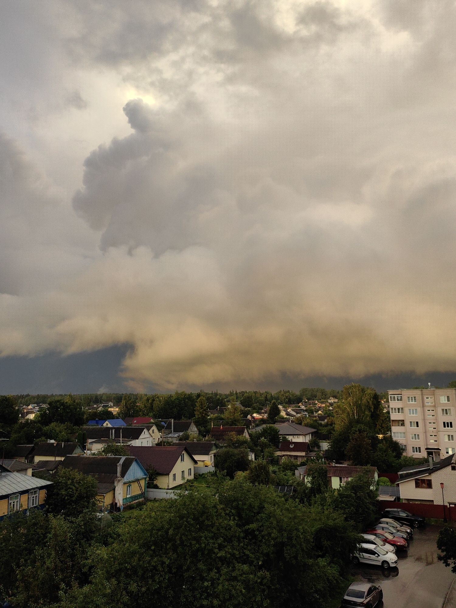 This aerial photograph depicts a suburban landscape in the city of Fanipal, Belarus, under a dramatic sky filled with ominous storm clouds. The lower portion of the image showcases a dense collection of residential houses, characterized by varied architectural styles and roof colors, interspersed with lush green trees and vegetation. A parking lot with several cars, including a white sedan, a dark sedan, and a red car, is visible to the right. Further to the right, a multi-story building with numerous windows stands out against the backdrop of the sky. The sky itself is a spectacle of contrasting light and dark, with bright, illuminated clouds juxtaposed against darker, storm-laden masses, suggesting an impending downpour or the aftermath of a storm. The overall impression is one of a tranquil residential area preparing for or experiencing inclement weather.