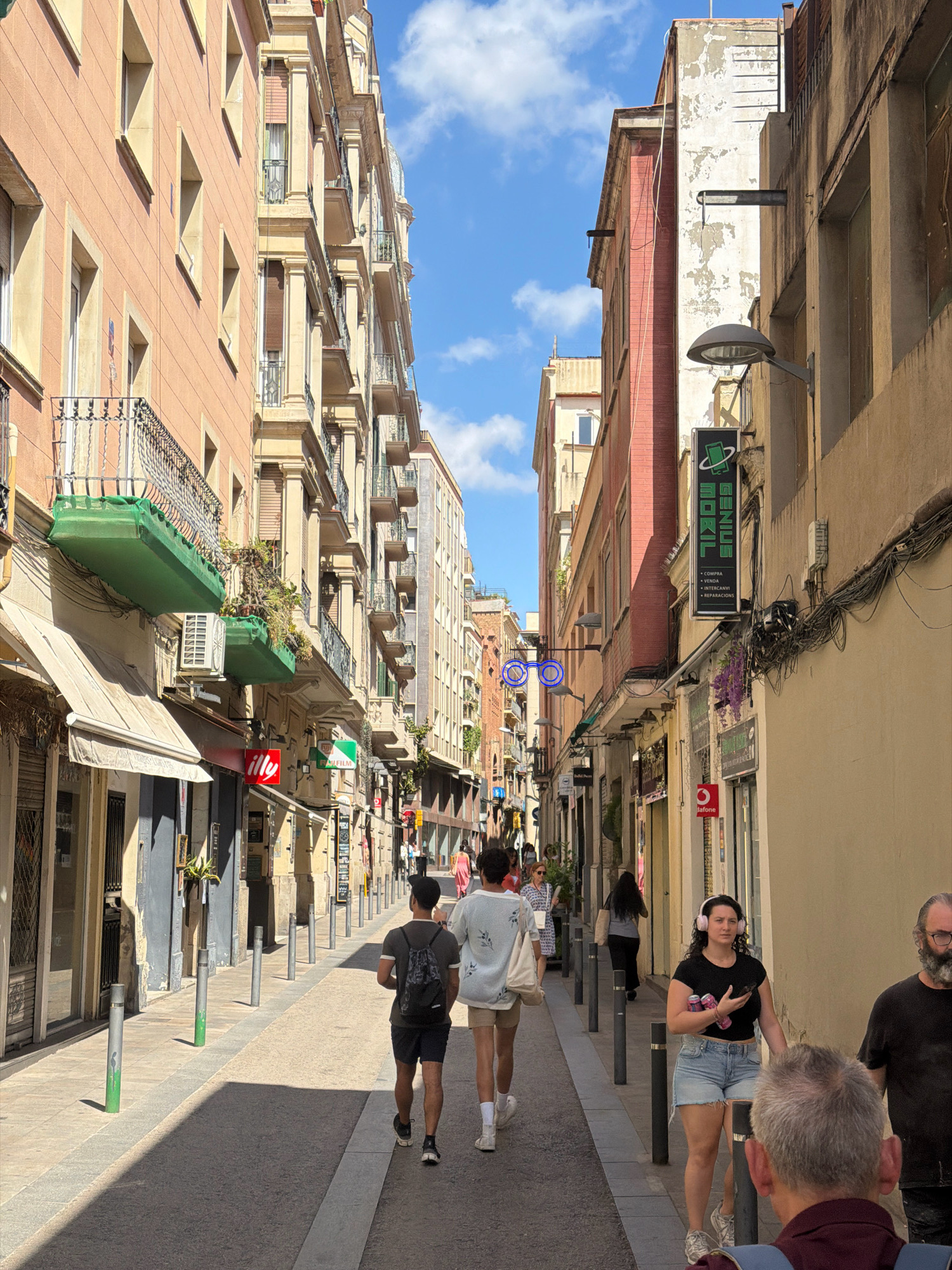 The image depicts a narrow, pedestrianized street in Barcelona, Spain, lined with multi-story buildings. The buildings exhibit varied architectural styles, with some featuring ornate balconies and classical detailing, while others have a more utilitarian appearance with peeling paint. The street is paved with asphalt and has a gray curb. Several cylindrical bollards, some with green painted bases, are spaced along the sidewalk.
On the left side of the street, shops with awnings are visible, one displaying an "illy" logo. Further down, a Fujifilm sign is mounted on a building. On the right side, a tall, vertical sign for "GENIUS MOBIL" is prominent, with smaller text below indicating "COMPRA • VENDA • INTERCANVI • REPARACIONS" (Buy, Sell, Exchange, Repairs).
Several people are present in the scene. In the foreground, two young men are walking away from the camera, one wearing a backpack and shorts, the other in a light-colored patterned shirt and shorts. To the right, a young woman with headphones on is walking towards the camera, wearing a black top and denim shorts, holding a smartphone and two cans. An older man with a beard is also visible in the right foreground, walking away from the camera. Other individuals can be seen further down the street, some walking and others standing.
The sky above is partly cloudy, with patches of blue visible. The buildings cast long shadows across the street, indicating it is daytime. The overall atmosphere suggests a sunny day in a bustling urban environment.