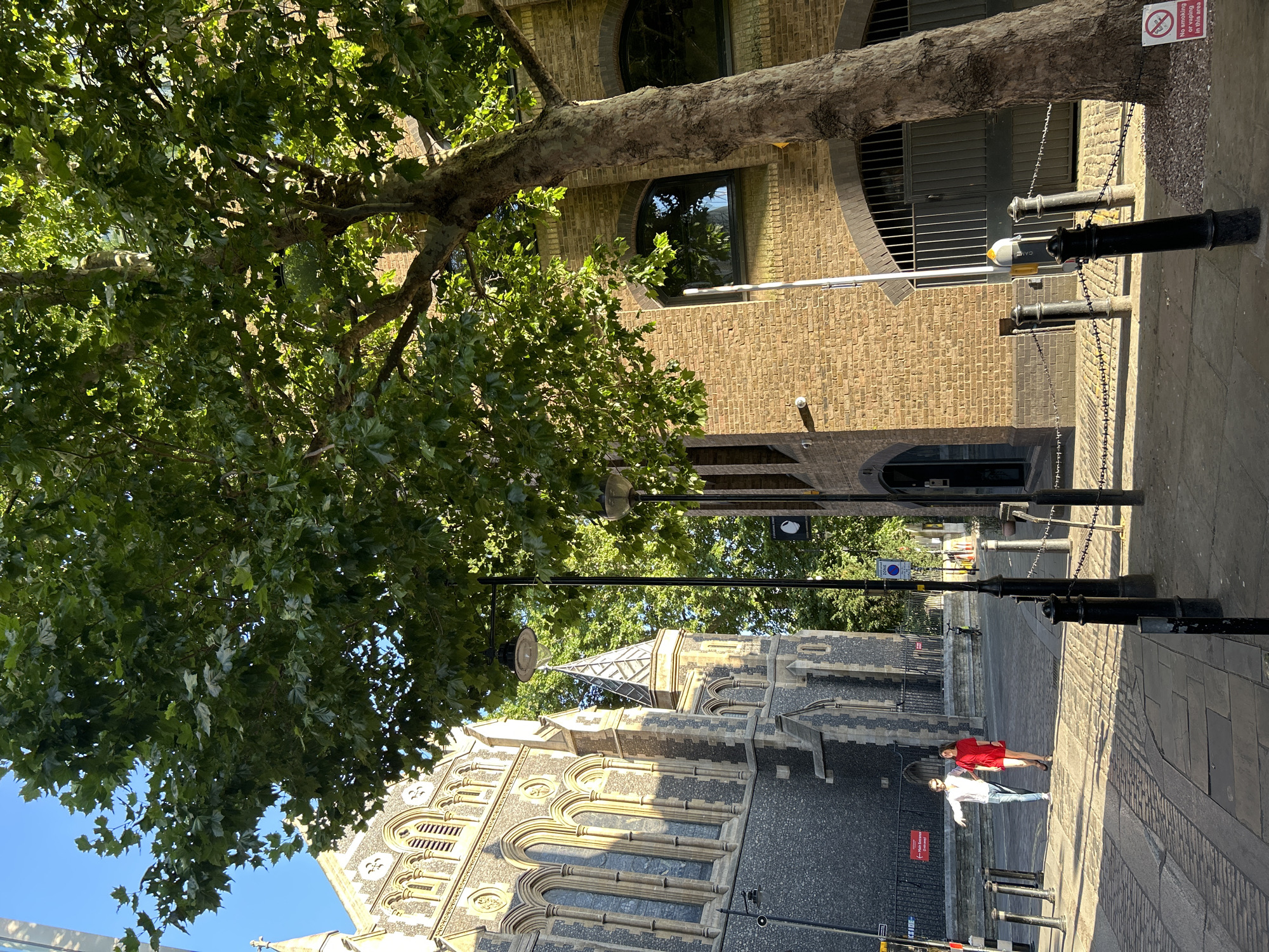 This is a street-level, medium shot photograph taken on a sunny day in London, United Kingdom. The scene depicts an urban environment with a mix of historical and industrial architecture. On the left is a large, imposing stone building with Gothic architectural features, likely a church, characterized by arched windows and intricate stonework. To the right, a building constructed of yellowish-brown brick with arched doorways and windows suggests an older industrial or commercial structure. A large tree with dense green foliage dominates the foreground and mid-ground on the right side of the image, casting dappled shadows on the brick building and sidewalk.
In the mid-ground, a man and a woman are walking side-by-side on a paved street. The man is wearing a light-colored long-sleeved shirt and jeans, and the woman is wearing a red dress. They are walking away from the camera, towards the left side of the frame. The street is made of cobblestones and flagstones, with bollards and chains lining the path. Several lampposts are visible, one with a distinctive ornate design. In the background, other buildings and trees can be seen, and the sky is clear blue. A "No Smoking or Vaping in this area" sign is visible on the lower right. The overall impression is a vibrant, sunlit street scene in a historic part of London.