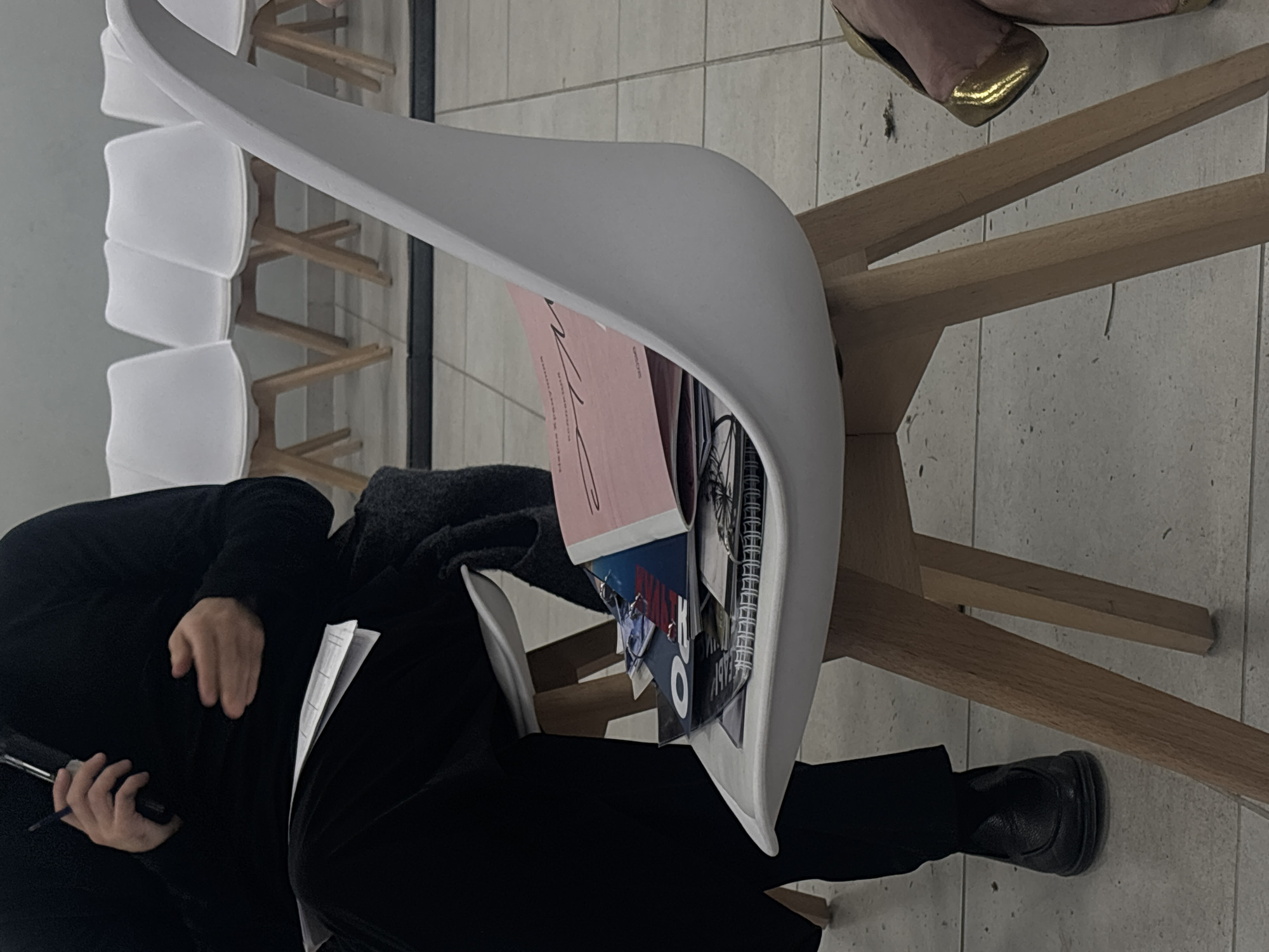 The image shows a person sitting on a white chair. The person is wearing black clothing and shoes. They are holding a pen and papers. Other chairs are stacked on the left. Some reading material is placed on the chair. The image was taken in the city of Perm, Russia.