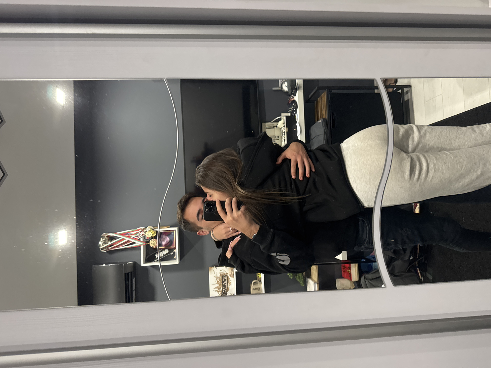 A young couple is embracing in a mirror selfie. The female is wearing a black hoodie and light gray sweatpants, while the male is in a black hoodie and dark jeans. They are facing each other in a room decorated with various items on shelves and walls, including a television, trophies, artwork, and personal belongings. The room has a dark gray wall. The location is inferred based on the assumption that they live in Sofia, Bulgaria. There is no definitive evidence to support this beyond the image itself.