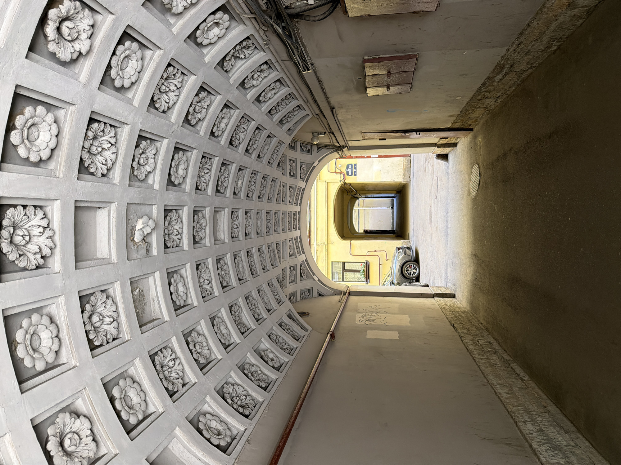 The image depicts a narrow architectural passageway in Saint Petersburg, Russia, oriented vertically, creating a dynamic perspective of a vaulted ceiling and an adjacent wall. The left side of the frame showcases an elaborately decorated, curved ceiling, characterized by a repetitive pattern of recessed square panels. Each panel contains an intricate white bas-relief sculpture, alternately depicting a detailed floral bloom and a leafy motif, all rendered in a classical style. The grey surface of this ornate ceiling shows signs of age, with some wear and minor scuffs.
In stark contrast, the right side of the passageway features a plain, unadorned wall and ceiling, painted in muted tones of grey and brown, indicating a more utilitarian function. Exposed electrical conduits and utility pipes run along the upper edge and side of this simple structure. Soft, indirect daylight illuminates the scene, suggesting either an overcast day or light filtering in from the opening ahead.
The passageway opens into a brighter area through an archway, revealing walls painted in a lighter yellow hue. Partially visible beyond the arch is a small, light-colored vehicle, possibly a compact car like a SMART car, parked in the distance. The ground of the passageway is a flat, grey surface with subtle textural variations. No people are present, and no discernible activities are taking place. The scene offers a blend of historical architectural grandeur and functional urban infrastructure, with no legible text visible within the frame.