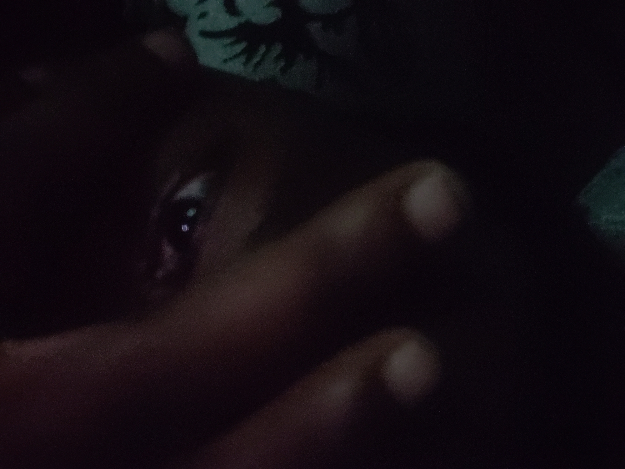 The image shows a close-up, low-light, and blurry view of a person's face, with their fingers obscuring parts of it. The subject's left eye is visible, reflecting light. A dark, possibly patterned fabric is partially visible in the background. The scene's location in Iganmu, Nigeria, is not visually discernible from the image itself.