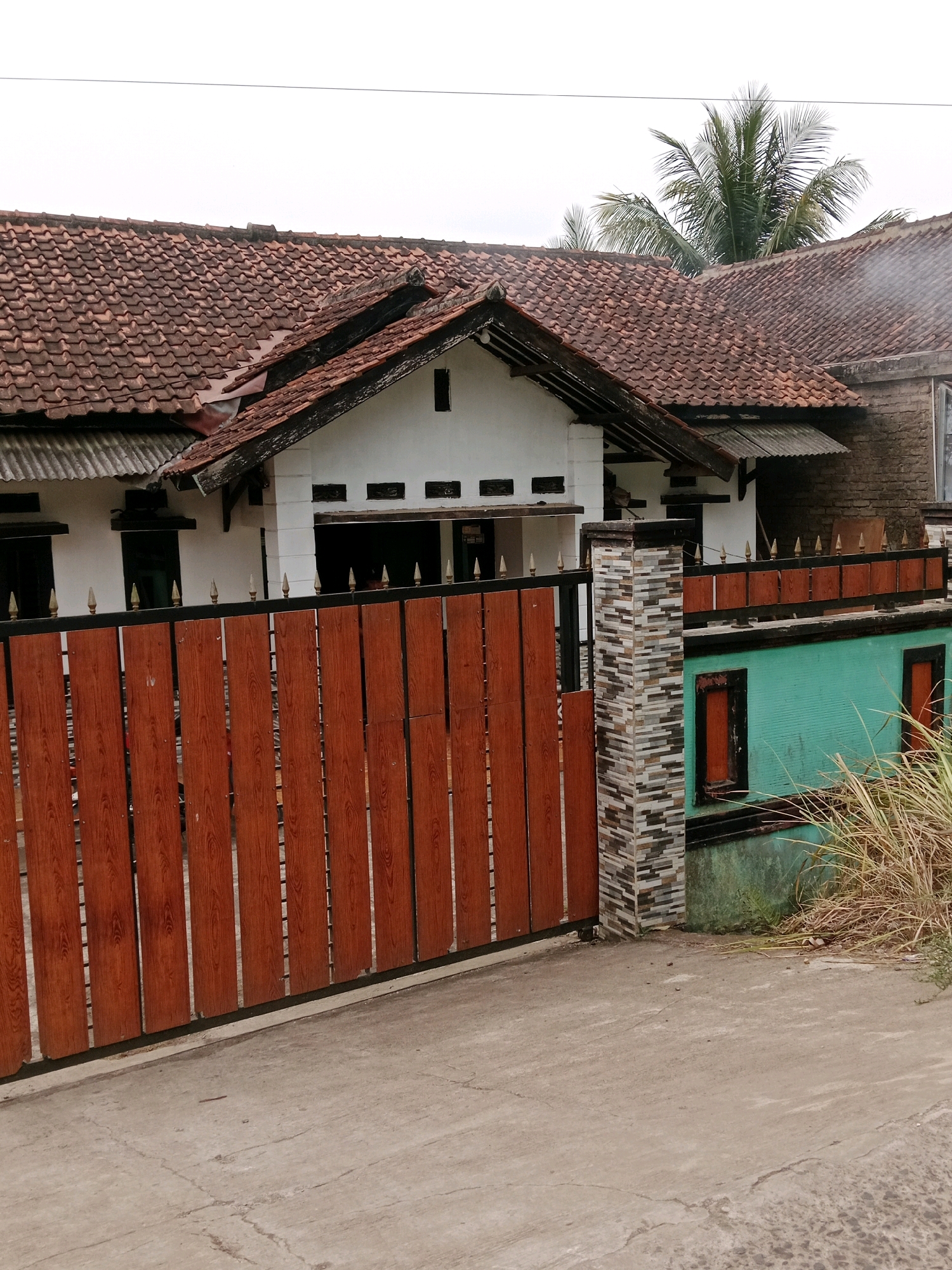 The image depicts the front exterior of a residential building in Cibinong Hilir, Indonesia. The house features a terracotta tile roof and white walls. A tall wooden gate with vertical planks is prominently displayed in the foreground, with decorative metal finials along the top. To the right of the gate, a section of the wall is painted a vibrant turquoise, incorporating decorative stone pillar accents. Dry, tall grass grows on the right side of the property, bordering the turquoise wall. The scene is captured on an overcast day, with a clear sky visible above the roofline. There are no people or vehicles present in the frame.