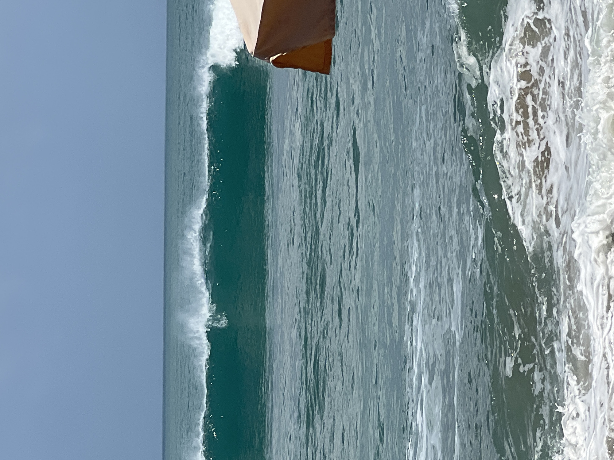 A medium shot captures a serene ocean scene in Kuta, Indonesia, under a clear, pale blue sky. The turquoise water is calm, with a large wave forming in the distance, its crest breaking into white foam. In the foreground, a beach umbrella, its canopy a muted tan and orange, partially juts into the frame from the right. The ocean stretches towards the horizon, meeting the sky in a seamless blend of blue. The composition is dominated by the tranquil blues and greens of the water and sky, with the umbrella providing a touch of man-made color.