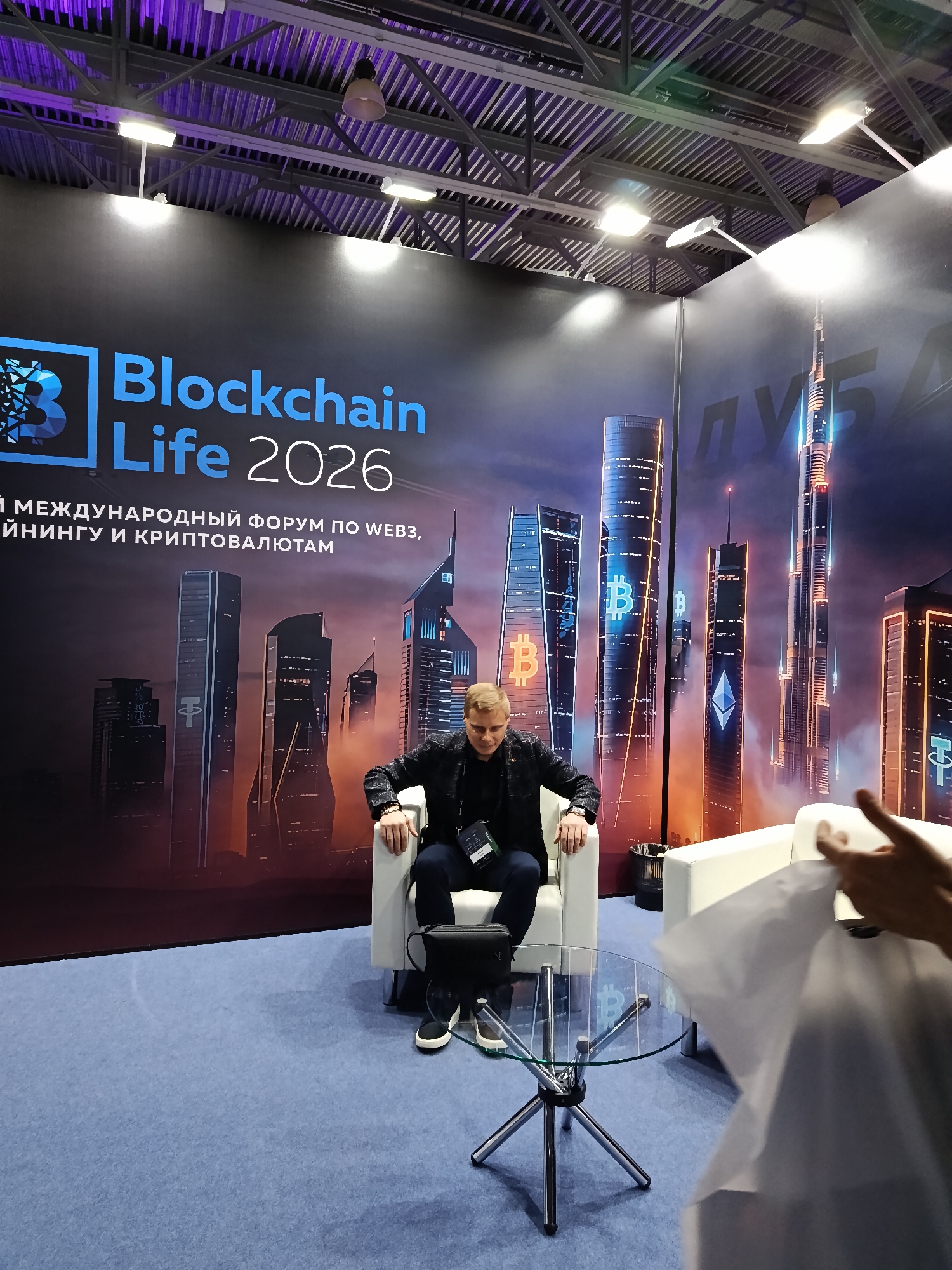 The image depicts a man sitting in a white armchair at a "Blockchain Life 2026" event. Behind him is a large backdrop featuring city skyscrapers with Bitcoin and Ethereum symbols. The backdrop includes the text "Blockchain Life 2026" and other text in a non-English language. In front of the man is a glass-topped table with a Bitcoin logo visible through the glass. A white sofa is to the right, partially obscured by a person and some white material, possibly a bag or cloth. The setting appears to be an exhibition or conference area with blue carpeting and a ceiling with industrial lighting and metal supports.
