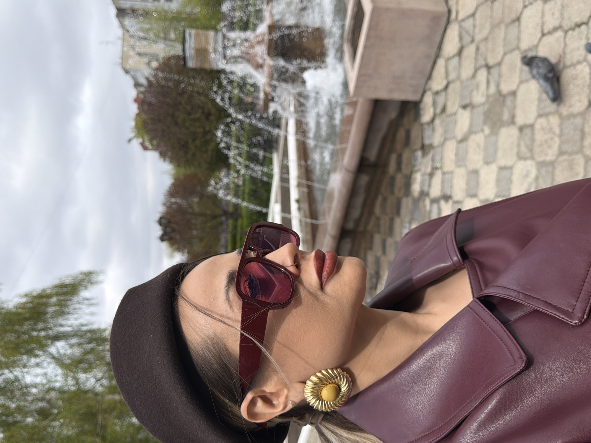 A woman wearing a brown beret, sunglasses, a maroon leather jacket, and a gold flower-shaped earring is positioned in front of a fountain with water spraying upwards. The fountain is located on a stone-paved surface. In the background, there are buildings and trees, and a pigeon is on the right side of the stone-paved surface. This location is in the city of Krasnoyarsk, Russia.