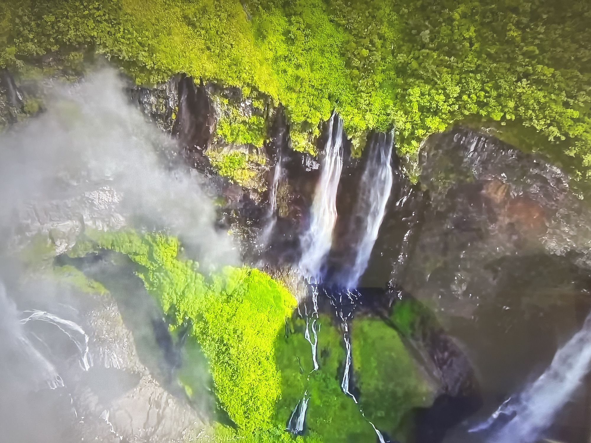 The image shows a view of a waterfall from above. The waterfall cascades over a cliff face that is covered in lush green vegetation. Mist is present around the waterfall. There is no indication of Basingstoke and Deane in the image. There are no people, subjects, objects, actions, or interactions to be described other than the waterfall and surrounding environment.