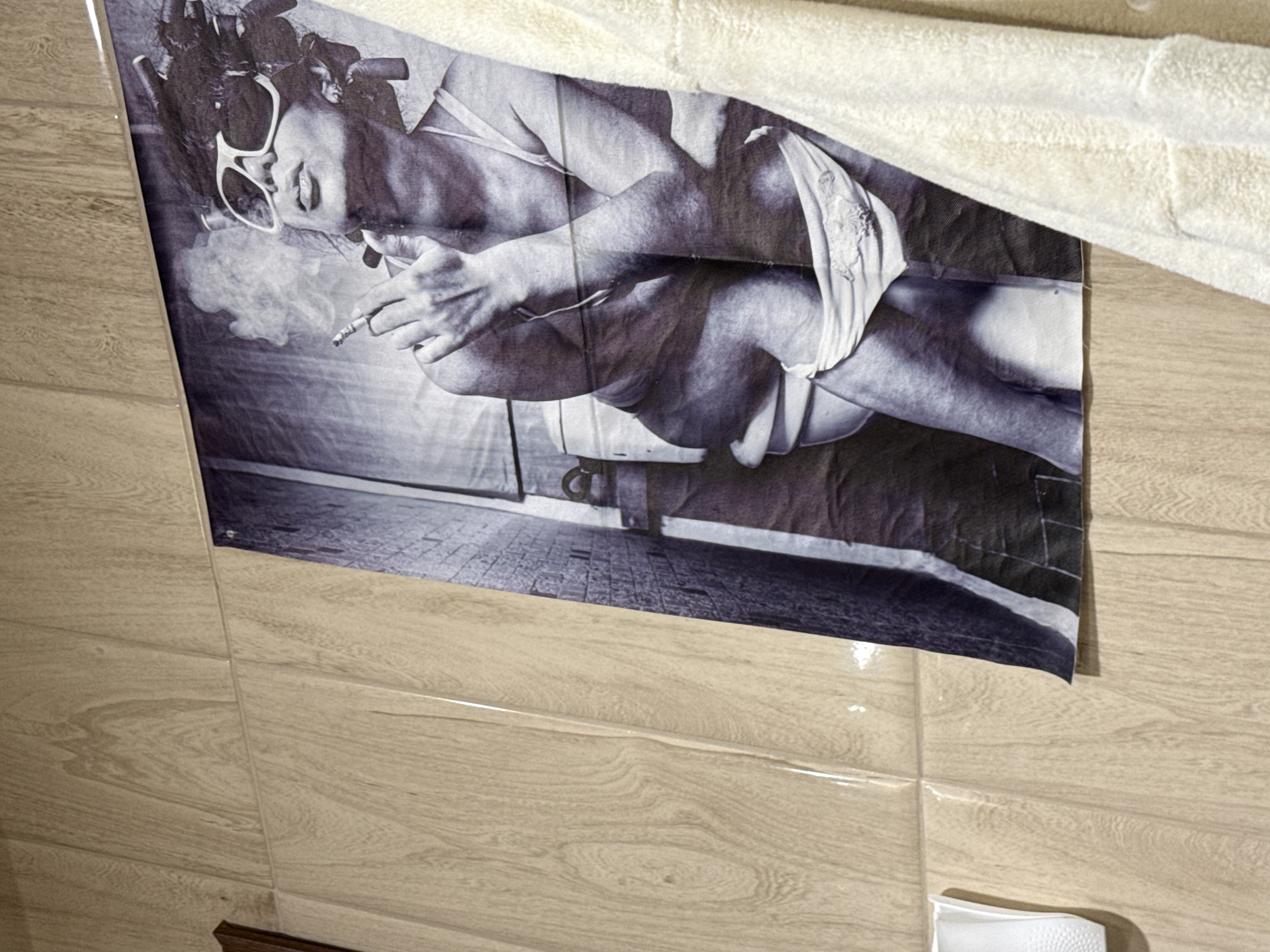 A black and white poster of a woman in sunglasses and hair rollers smoking a cigarette is hanging on a wall with wood-patterned tiles. A white towel obscures part of the top of the poster. A small white container is visible near the bottom right. There is no discernible evidence to place the scene in Iarebichna, Bulgaria.