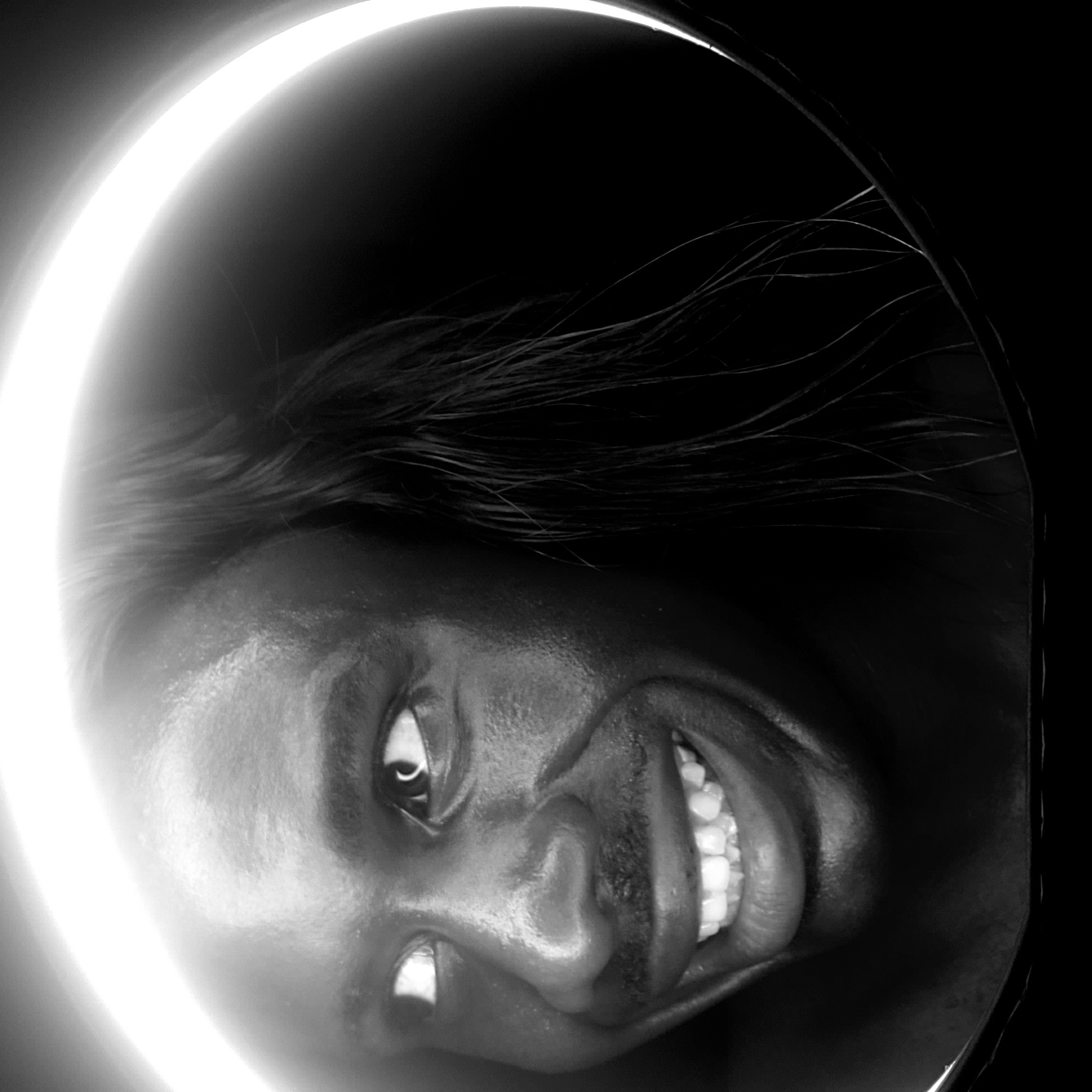 A black and white photograph, rotated counter-clockwise, shows a person with dark skin facing the camera and smiling, teeth visible. The person has short, dark hair. A ring of bright light surrounds the person's head. The background is dark. The location is not discernible from the image.