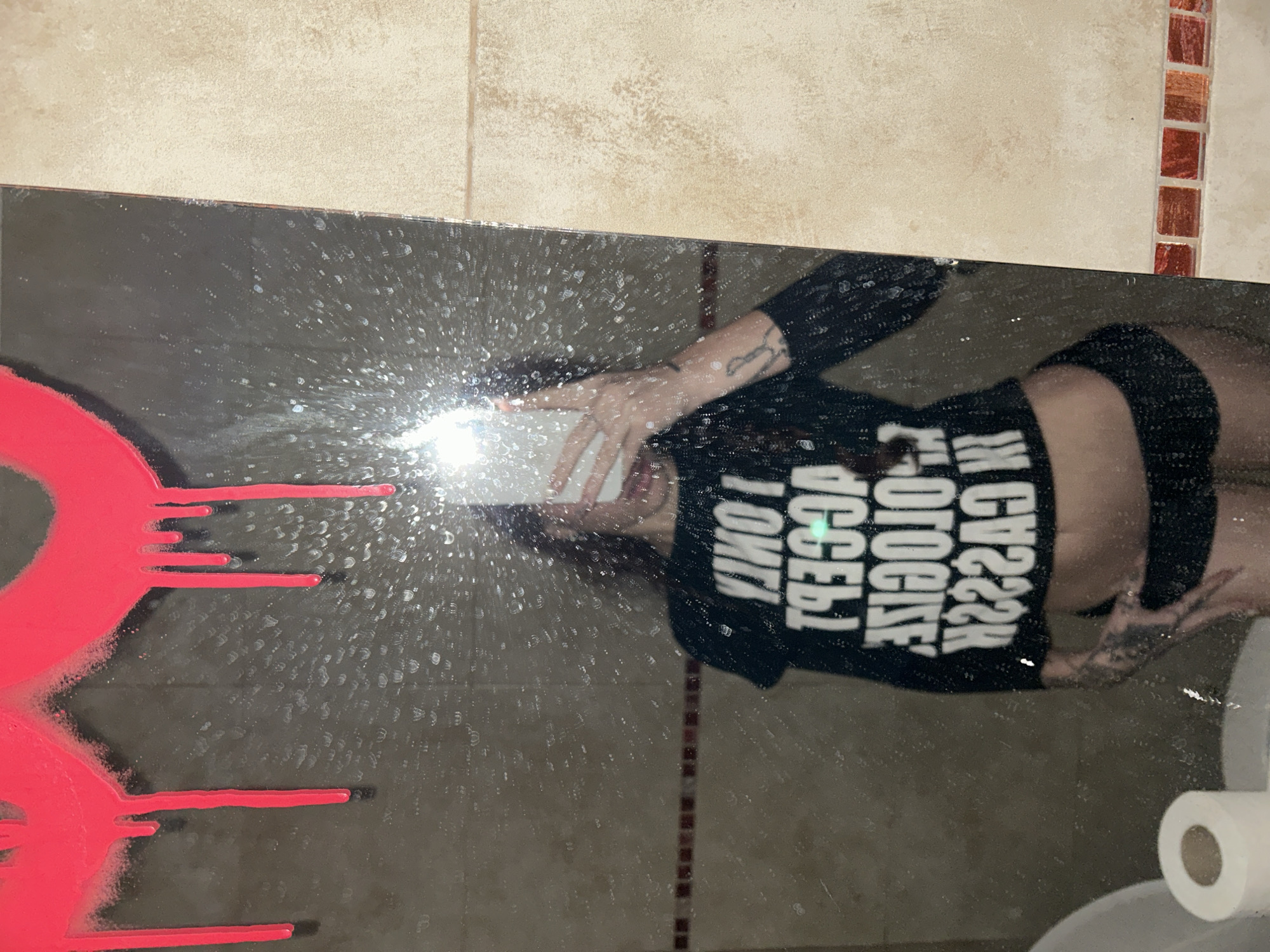 A selfie is taken in a dirty bathroom mirror. On the left side of the mirror, there is red paint dripping. A person in a black long-sleeve shirt with black shorts is reflected in the mirror, holding a phone. The words on the shirt appear reversed, "I ONLY ACCEPT APOLOGIES IN CASH". The lower-right corner shows a toilet and a roll of toilet paper. The walls have beige tiles, with a vertical strip of brown and beige patterned tiles. The location is inferred to be in Buenos Aires, Argentina.