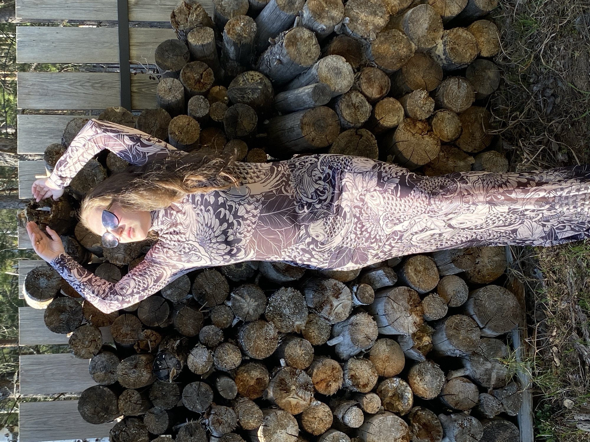 A woman wearing sunglasses and a patterned, long-sleeved dress is lying on a stack of cut logs, likely firewood. The logs are stacked within a rough frame, with some vegetation visible nearby. The location is presumably rural or at least adjacent to a wooded area, but there is no direct evidence indicating that the location is Markovo, Russia.