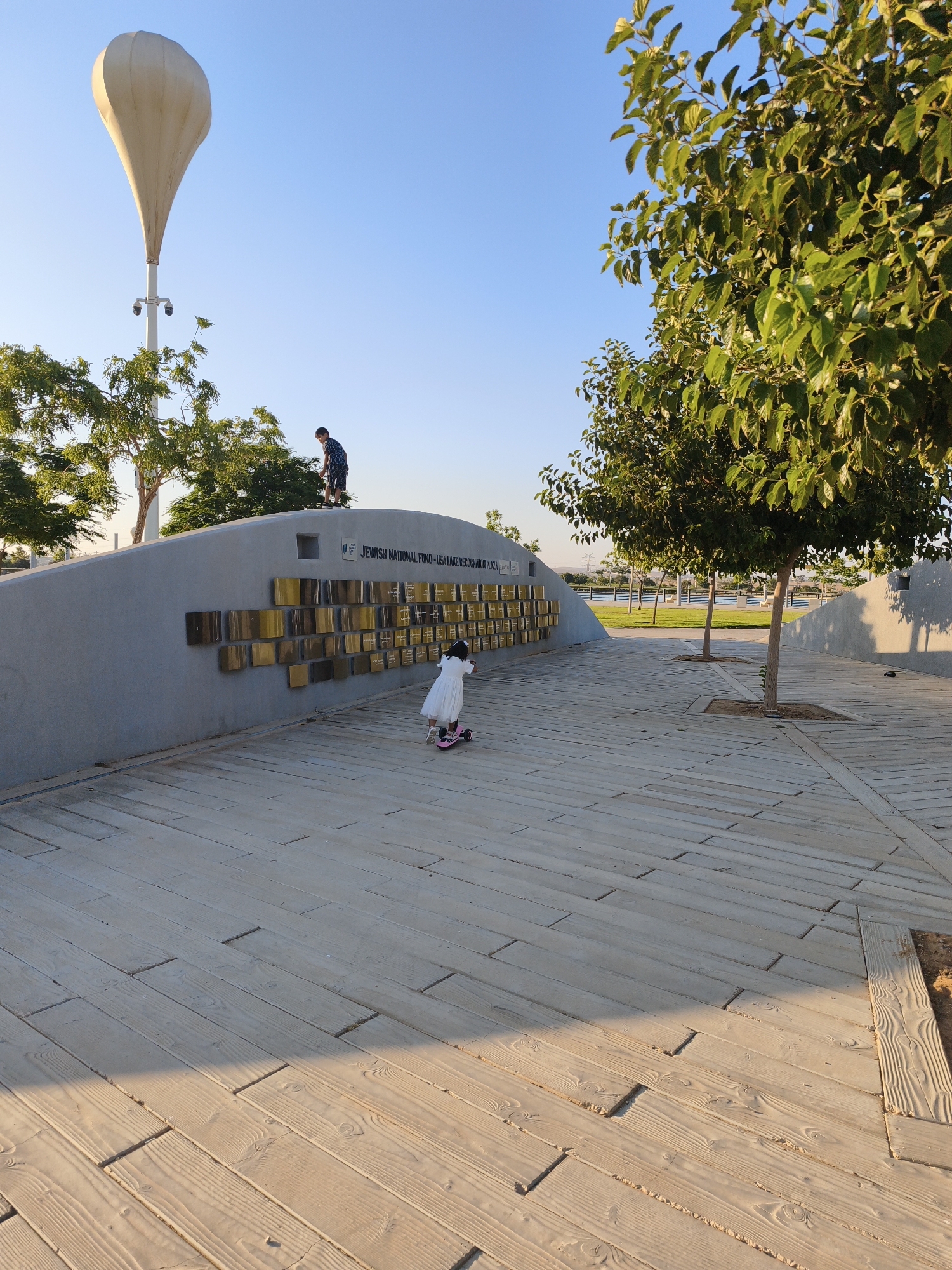 In Be'er Sheva, Israel, on a sunny day, a young boy stands on a curved concrete wall overlooking a wooden promenade. Beside him, a young girl on a scooter rides away from the wall. The wall features a plaque inscribed with "JEWISH NATIONAL FUND - USA LAKE RECOGNITION". Rows of golden plaques are embedded in the wall, likely commemorating donors. In the background, a large, light-colored balloon-like structure sits atop a pole, flanked by trees and a clear blue sky. The wooden promenade, made of light-colored planks, leads away from the wall, with trees planted at intervals. The scene suggests a public park or memorial site.