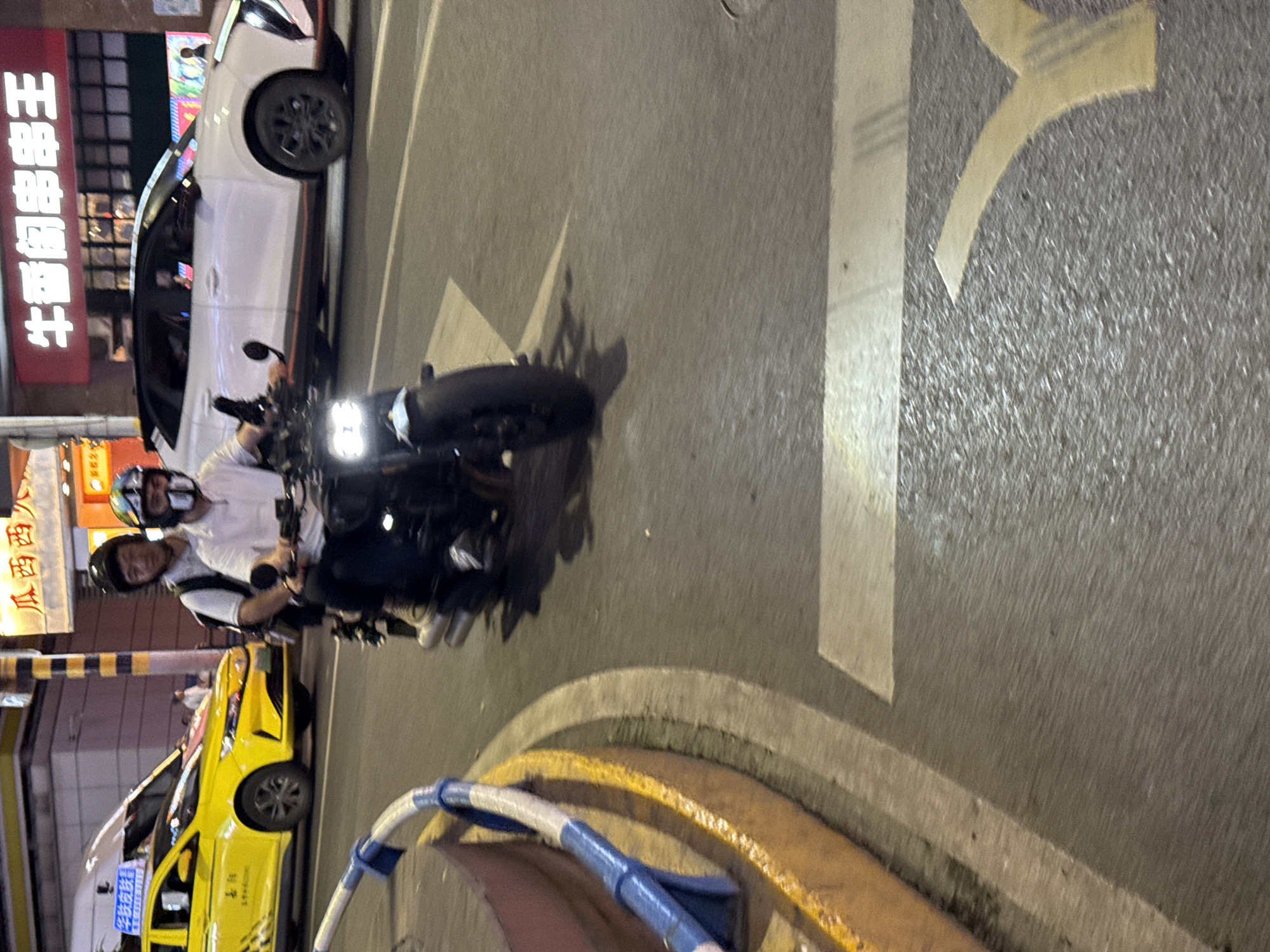 The image captures a nighttime street scene in a city, likely Yuzhong District, China, as indicated by the context and visible Chinese text. The perspective is tilted, showcasing the road, a motorcycle, and various vehicles alongside commercial buildings.
In the center, two individuals are riding a black motorcycle. The driver, wearing a white shirt and a black helmet, is positioned in front of a passenger who wears a black shirt and a black helmet. The motorcycle's bright, rectangular LED headlight is prominently illuminated.
Adjacent to the motorcycle, a white car with dark wheels is visible, appearing to be parked or moving slowly. Further down the street, a yellow taxi with checkered patterns on its side is also present. The road surface features clear white lines and a distinctive yellow arrow marking. A low concrete barrier, partly covered with blue and white striped material, curves along the right side of the street.
Building facades line the street, adorned with illuminated signs. A large red sign prominently displays the Chinese characters "王哥串串香" (Wang Ge Chuan Chuan Xiang), indicating a restaurant or food stall. Other less legible Chinese characters are visible on signs in the background. The overall illumination from streetlights and shop signs confirms the nighttime setting. The road appears dry. The people on the motorcycle are wearing helmets and appear focused on their ride.