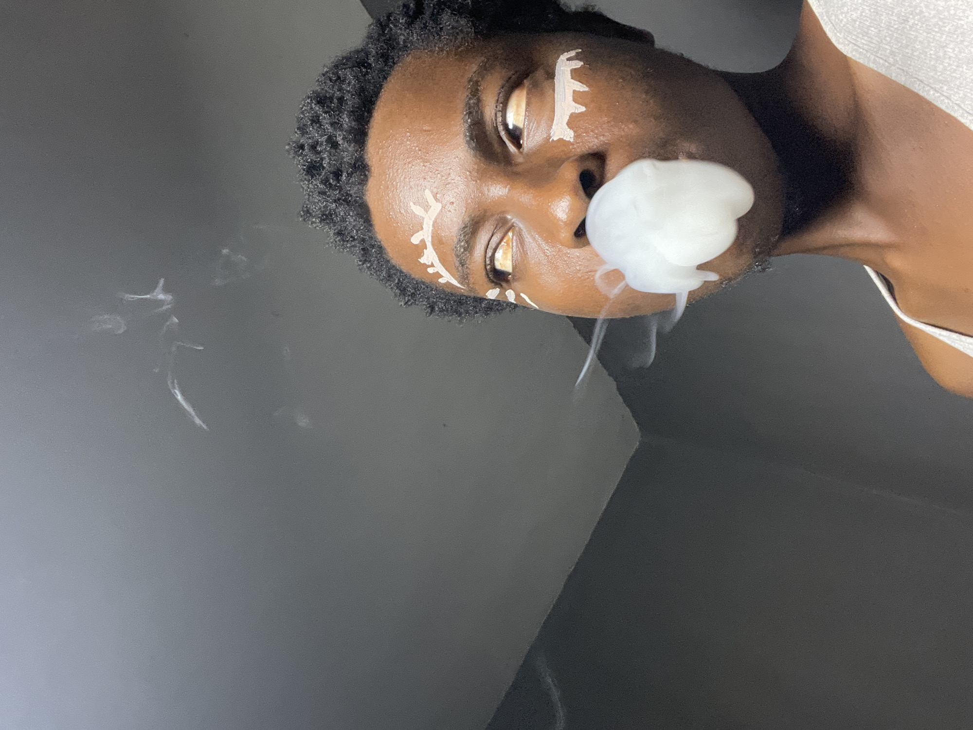 A person with dark skin and short, dark, curly hair is looking upwards and to the left, exhaling a large cloud of white smoke from their mouth. They have white markings on their face, resembling stylized eyebrows and markings under their eyes. The person is wearing a light-colored, ribbed tank top. The background is a dark gray wall, with a lighter gray ceiling at an angle in the upper left corner. Wisps of smoke are visible in the air. This scene is located in Nkpor, Nigeria.