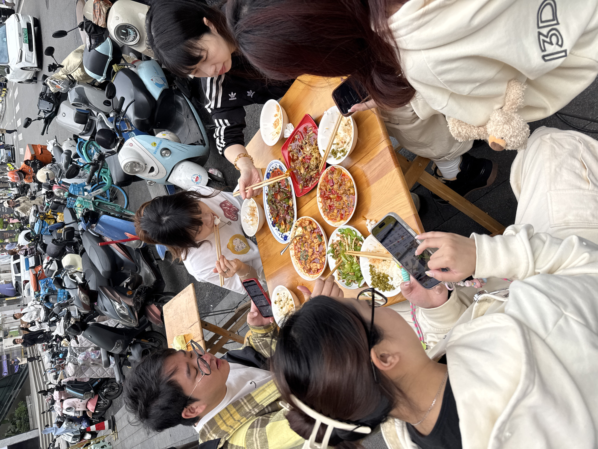 A group of five people are sitting around a wooden table covered with multiple dishes of food and individual bowls of rice. They appear to be eating a meal outdoors, near a row of parked motorbikes and scooters. The individuals are of Asian descent and are dressed in casual clothing. Some are using chopsticks to pick at the food, while others are holding and using smartphones. A few of the phones display photos of the food on the table. A white car is parked in the background along with a display of goods in the background.
The image was taken near the motorbikes near the food stalls in the Jinjiang District of China.