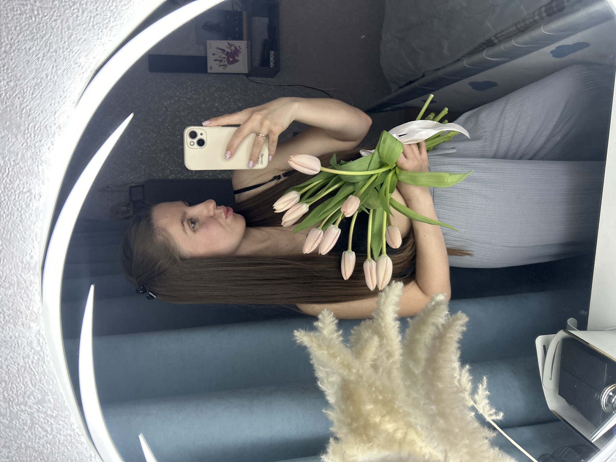 A young woman is reflected in a ring light mirror while taking a selfie with her smartphone. She is holding a bouquet of light pink tulips with green stems and leaves in her left hand. She has long brown hair, light skin, and is wearing a black top. She is wearing a gold ring on her left hand. Her nails are painted with a nude color and have small multicolored polka dots. The phone case is a light beige color.
In the background, reflected in the mirror, are various objects including a white cabinet, a bed with blue and white covers, and a box. In the foreground, a white feathery plant is partially visible. It is unclear if this image was taken in the city of Klin, Russia.