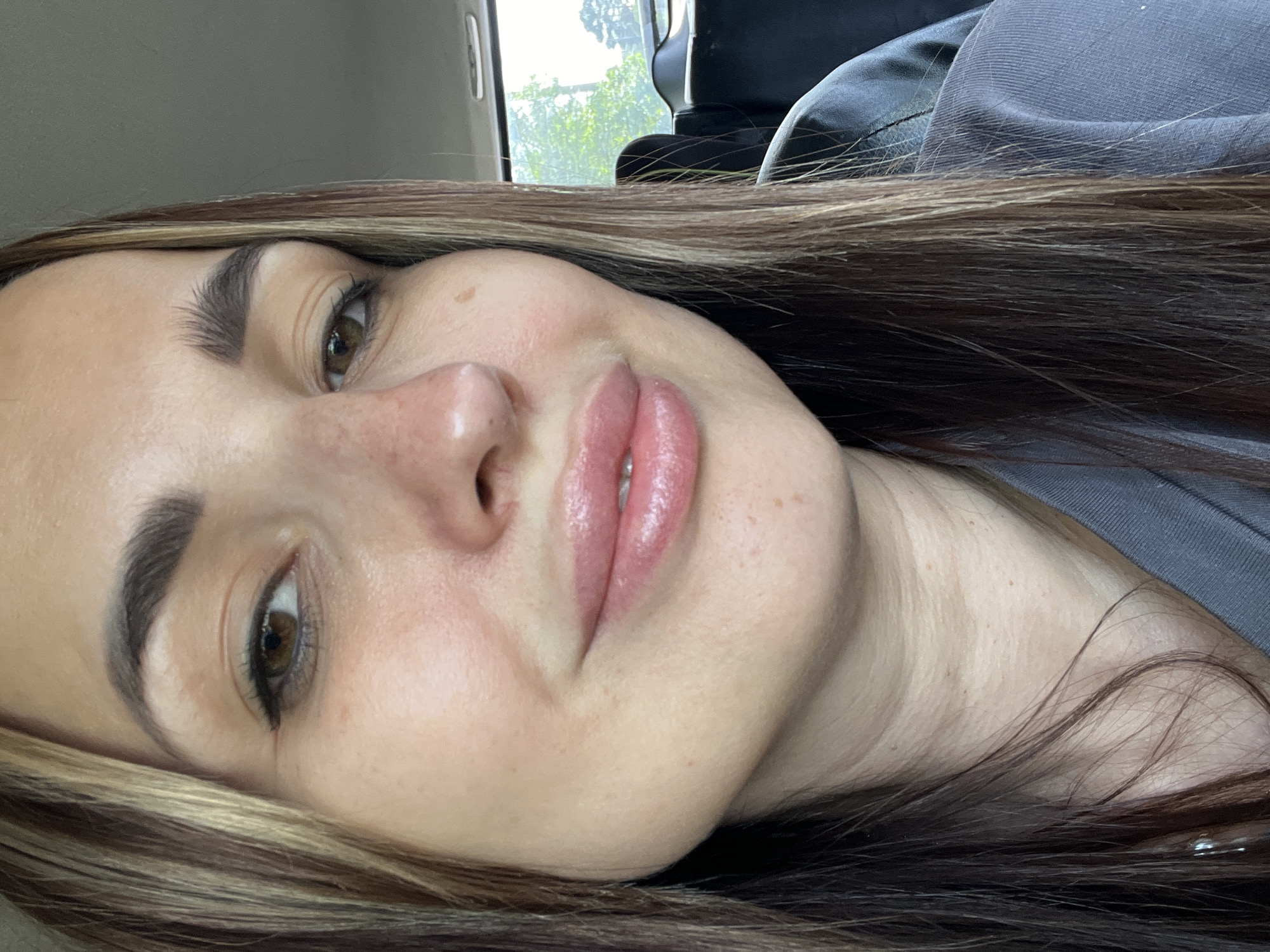 An over-the-shoulder, medium close-up shot shows a woman in her 20s with shoulder-length brown hair with blonde streaks. She is wearing a grey t-shirt and has makeup on her face, including eyeliner and lip gloss. The woman is looking at the camera and smiling slightly. The background is blurry and appears to be the interior of a car, with trees visible through the rear window. The lighting is natural and soft.