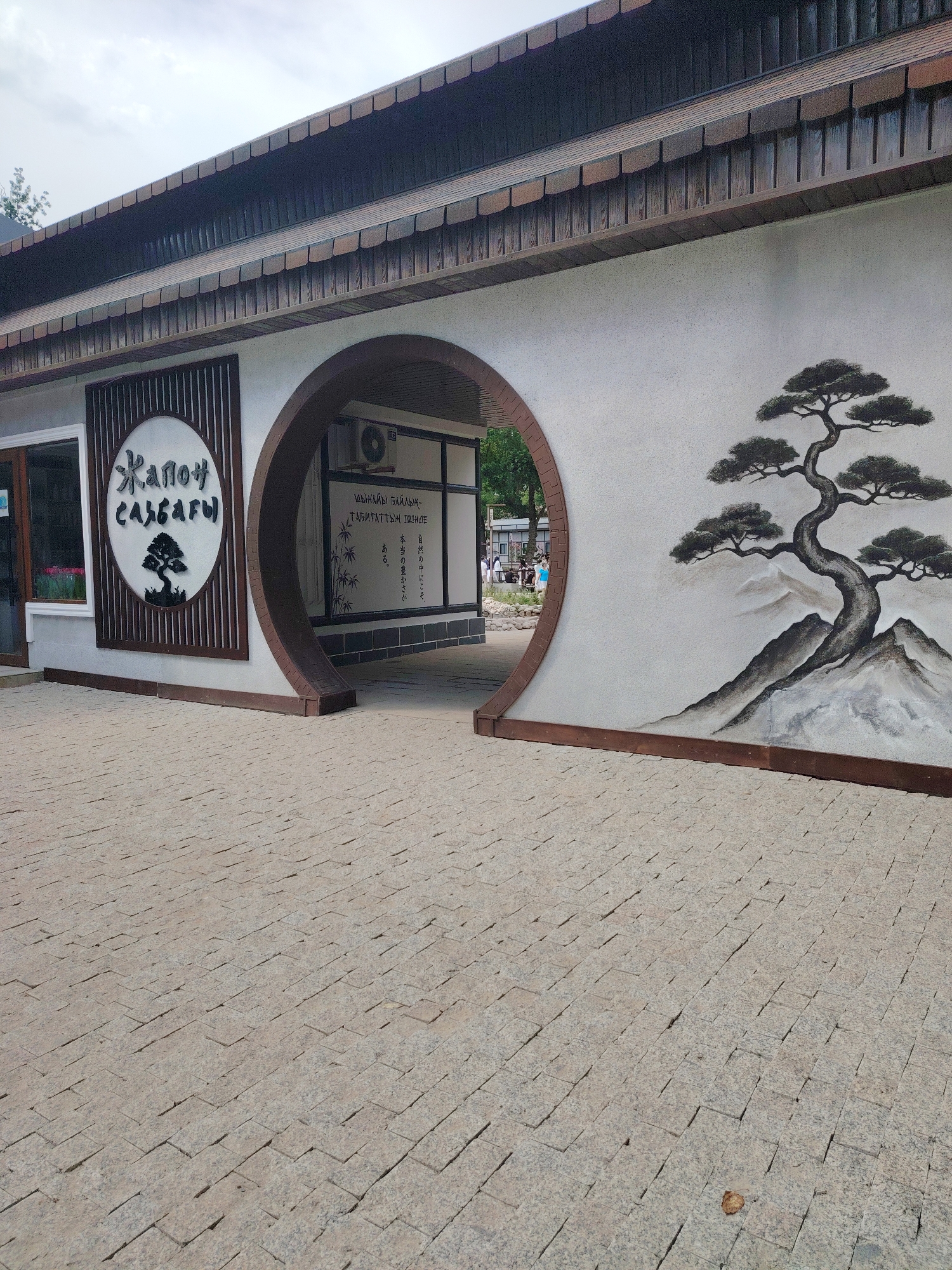 This media file shows an outdoor view of a building with a Japanese-inspired architectural style in Shymkent, Kazakhstan. The building features a white stucco exterior with a dark, sloped roof and a large, circular archway in the center, revealing a glimpse of an interior space. To the left of the archway, a sign with the text "Жапон САЛБАРЫ" is prominently displayed within a decorative frame, alongside an image of a stylized tree. Further to the right, on the wall, is a mural of a bonsai tree on a mountain landscape. Inside the archway, an air conditioning unit is visible. The ground in front of the building is paved with light-colored rectangular stones, creating a textured walkway. The overall atmosphere suggests a serene and culturally themed establishment. The sky appears overcast, indicating it might be a cloudy day. The time of day is difficult to determine precisely due to the lighting conditions, but it appears to be daytime.