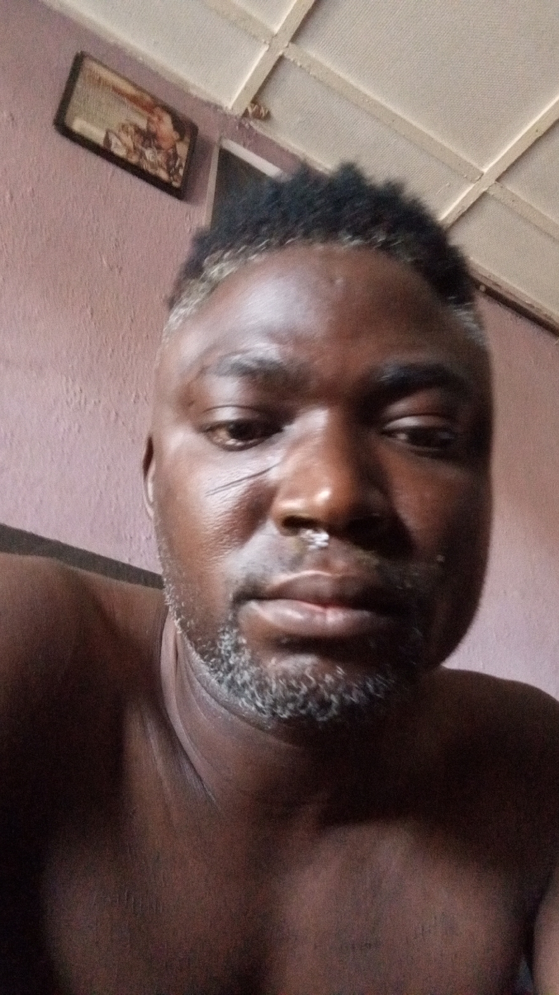 A close-up, eye-level selfie captures a dark-skinned man looking slightly downwards towards the camera. He is bare-chested, revealing his shoulders and upper chest. His short, curly hair is dark with noticeable streaks of grey or blonde, particularly on the sides. Sparse grey facial hair or stubble covers his chin and jawline. A distinct, slightly curved linear scar is visible on his right cheek, extending from near his eye towards his nose. There appears to be a white, somewhat powdery substance or object on his nose, close to the right nostril. His expression is neutral to slightly serious.
The background features a reddish-pink textured wall. In the upper left, a framed picture hangs diagonally, its contents indistinct due to blur. Above the man's head, a section of a light-colored, possibly suspended or tiled ceiling with a grid pattern is visible. A sliver of what appears to be a screen or monitor is partially seen to the right of his head. The indoor setting has subdued lighting. No text is discernible in the image.