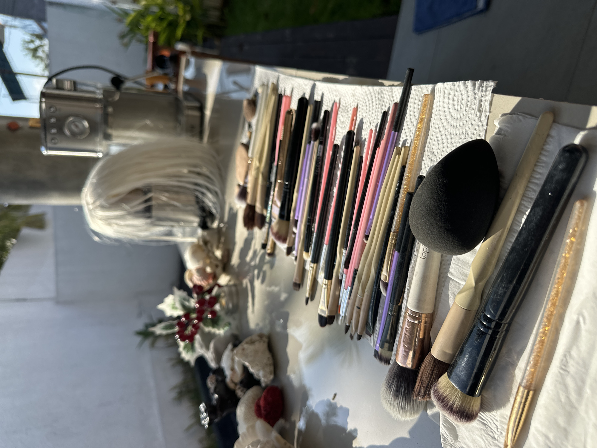 An outdoor scene is depicted in North Kuta, Indonesia, with a close-up view of makeup brushes arranged on a white paper towel. The brushes have varying handle colors including black, beige, rose gold, and purple. Some brushes are thicker with fluffy bristles, while others are thinner and more precise. In the foreground, a black makeup sponge with a rounded shape is prominent. The background shows a table with several items, including what appears to be a silver coffee maker, a white wig on a mannequin head, and decorative items with red berries and white leaves. The setting appears to be a patio or balcony, with sunlight casting shadows.