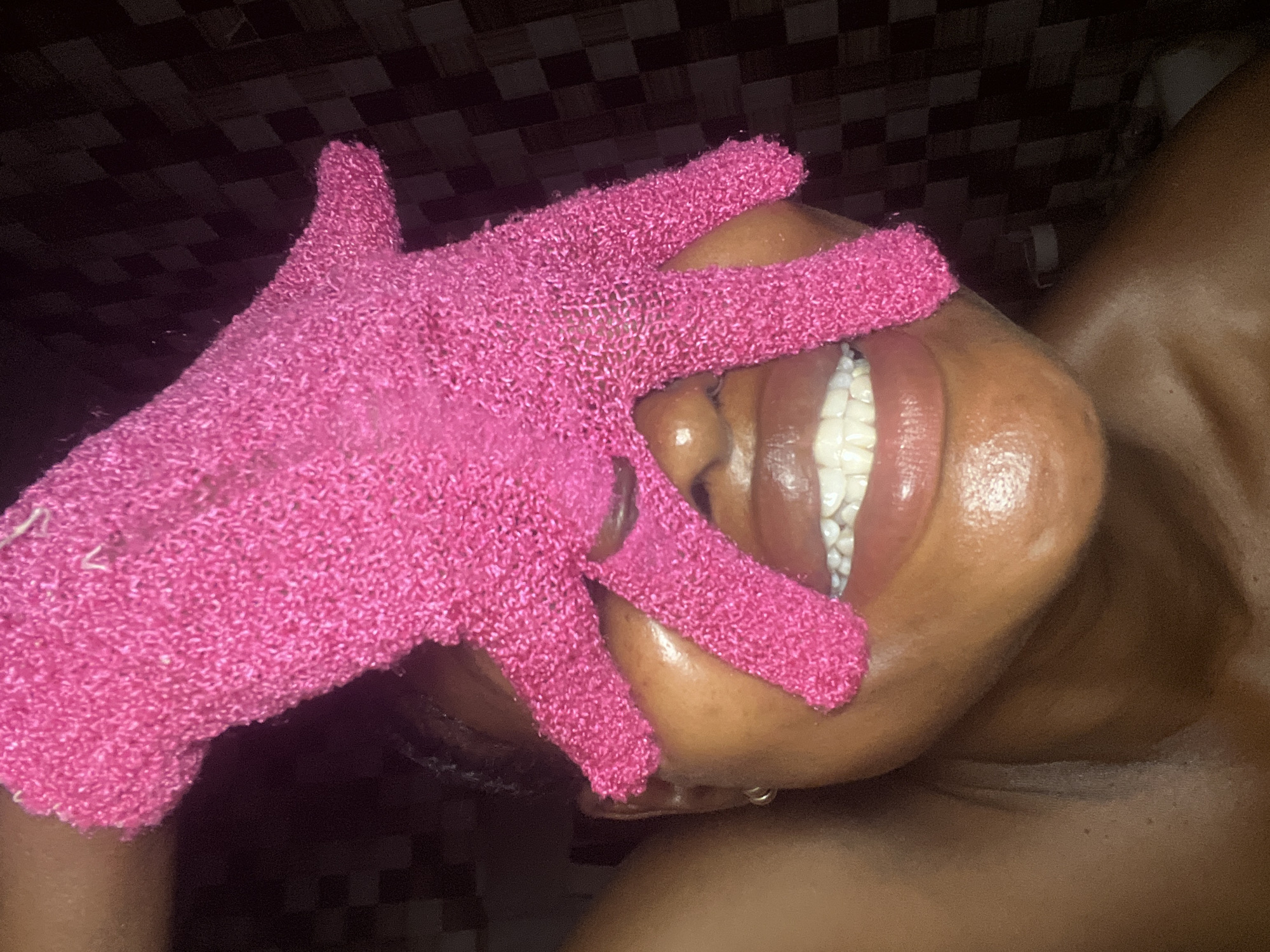 The image shows a close-up of a person's face, partially obscured by a bright pink exfoliating glove. The person's mouth is open, revealing white teeth and glossy lips. The skin appears to be moisturized and glowing. The background consists of a dark, checkered pattern, suggesting a bathroom setting, possibly in Ado, Nigeria. The lighting is dim, highlighting the subject's face and the texture of the glove.