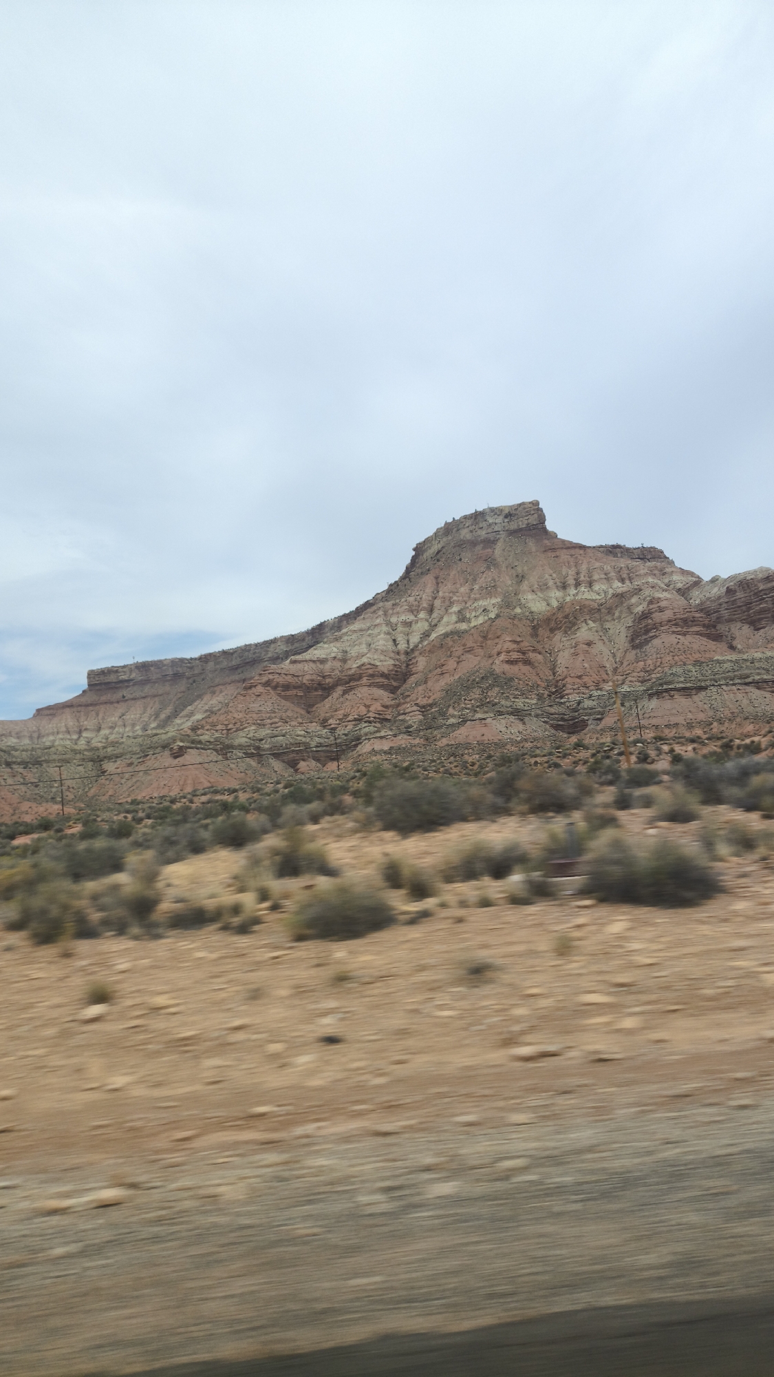 The image depicts a wide shot of a desert landscape with a prominent, layered mountain in the background. The mountain is characterized by distinct bands of red, white, and light brown rock. The foreground consists of dry, sandy terrain dotted with sparse, green desert shrubs. There are utility poles visible on the slopes of the mountain and in the mid-ground. The sky is overcast with a pale, cloudy appearance. The overall impression is of a natural, arid environment. The image is likely taken from a moving vehicle, as indicated by the motion blur in the foreground. The location is identified as being in the city of Virgin, United States of America.