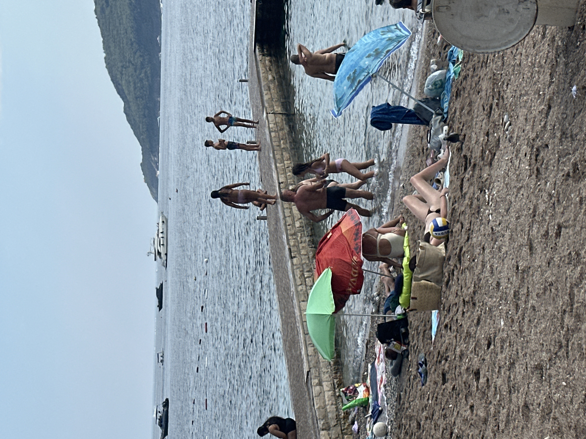 The image captures a sunny day at a pebbly beach in Budva, Montenegro, with a stone pier extending into the calm, blue sea. Several people are enjoying the water and sun. On the pier, a group of individuals, including a woman and three young men, are standing or walking. Near the shore, closer to the viewer, several people are relaxing on towels under umbrellas. A woman in a bikini is lying on the sand, and other individuals are wading in the shallow water or sitting near the water's edge. Beach accessories like towels, bags, a volleyball, and a ball are scattered on the beach. In the background, several boats and yachts are visible on the sea, and a lush green hillside forms the horizon. The overall scene depicts a typical summer day at a popular beach resort.