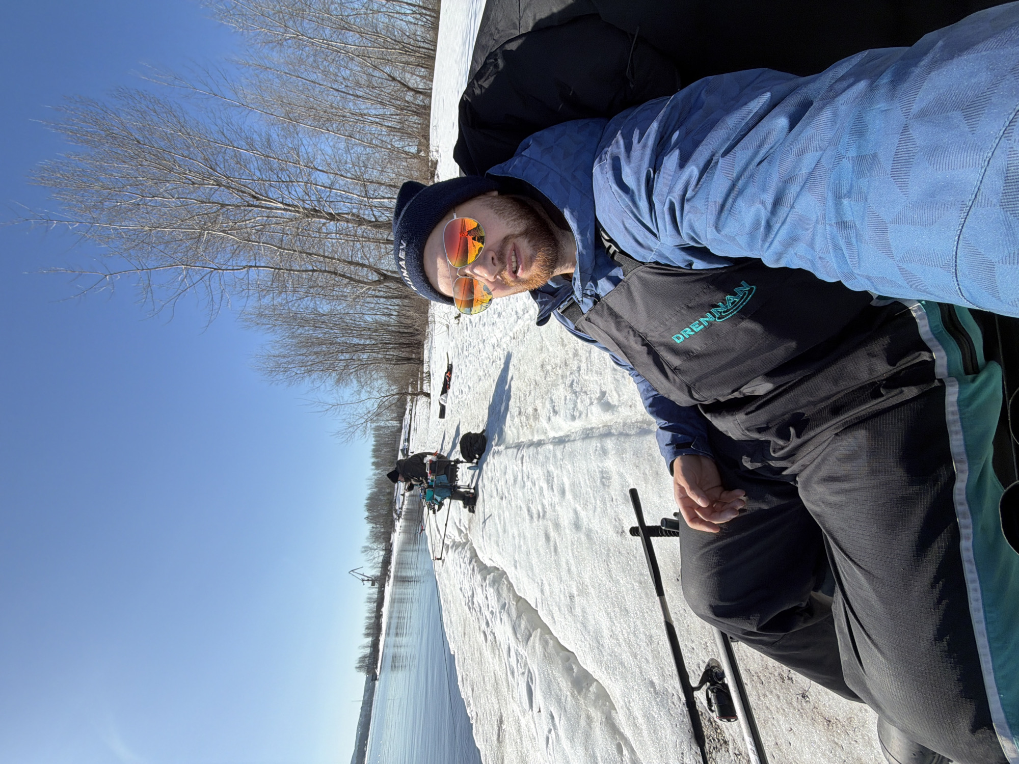 The image shows a man posing for a selfie on a snowy shoreline near a body of water, likely during an ice fishing trip in or around Chaikovsky, Russia. The man is wearing a blue jacket, black overalls with a "DRENNAN" logo, a dark-colored beanie, and mirrored sunglasses. He is seated, holding a fishing rod. In the background, another person is ice fishing, positioned near other fishing equipment on the snow-covered ice. The background includes bare trees and a clear blue sky. The presence of snow and ice suggests a winter season. A crane-like structure is visible on the opposite bank of the body of water.