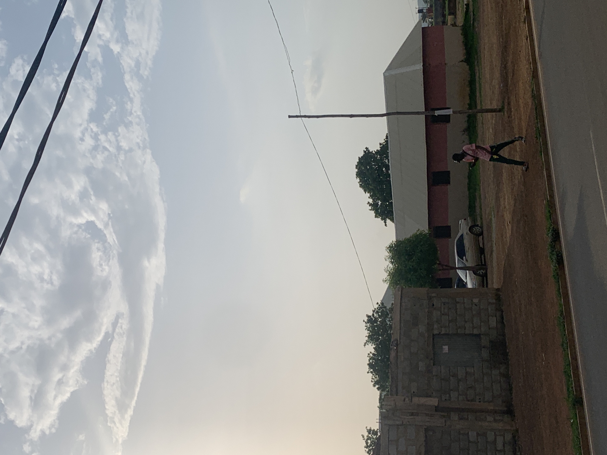 The image depicts an outdoor scene in Sabojibi, Nigeria. The sky is partially cloudy with electrical wires visible on the left side. A person wearing a pink shirt and dark pants is walking on the right side of the image, adjacent to a road. Behind the person is a white car parked near a building that is partially painted in dark red. In the foreground on the right side is a brick structure with a partially visible gray door. A wooden pole connected to wires extends across the sky.
