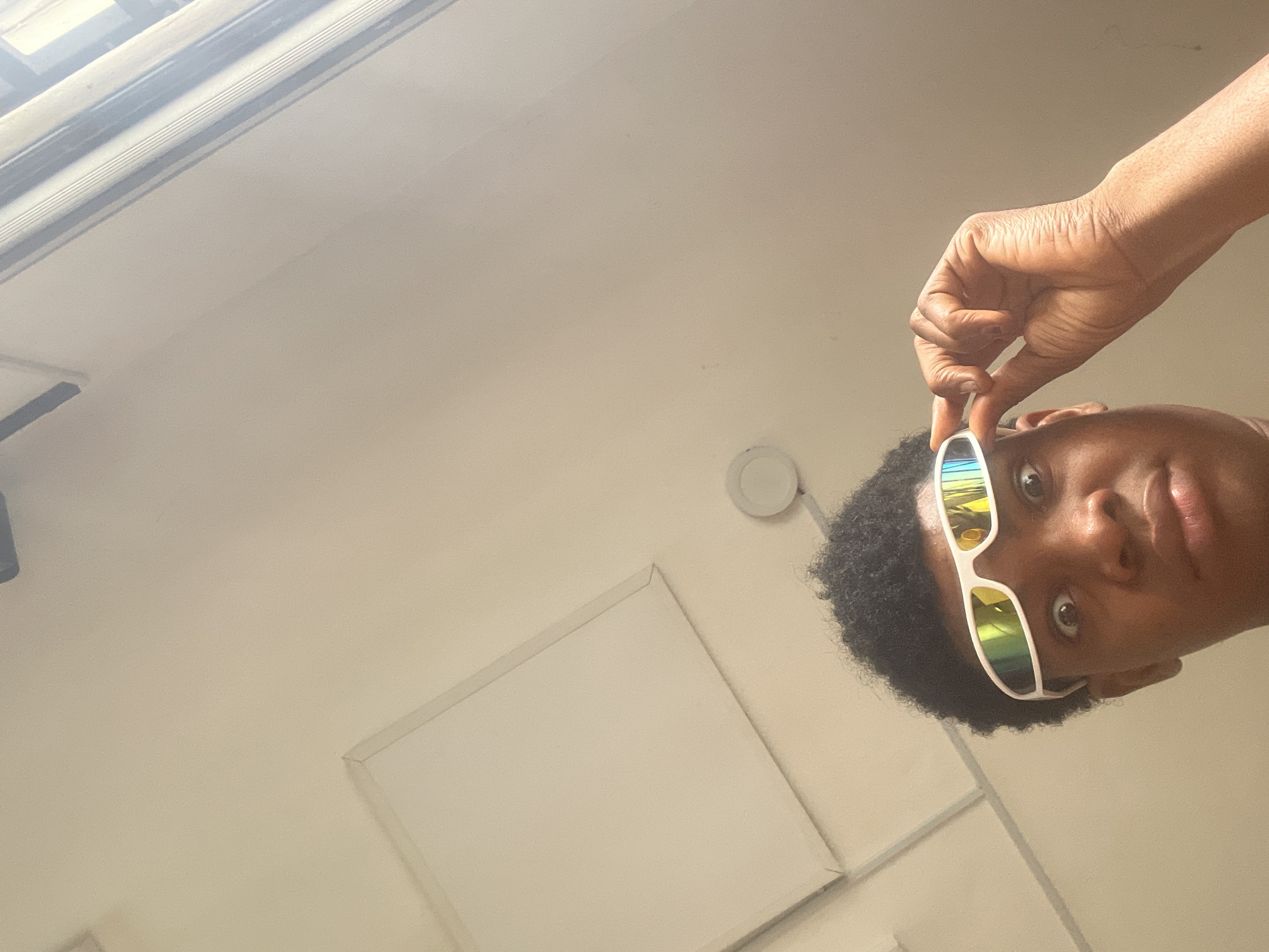 In Ado, Nigeria, a young person with dark, curly hair is looking up at the camera. They are wearing white-framed sunglasses with reflective, rainbow-colored lenses. One hand is raised, holding the sunglasses by the bridge as if adjusting them. The background is a neutral-colored wall and ceiling, with the edge of a window or light fixture visible at the top right. The overall impression is a casual, indoor setting.