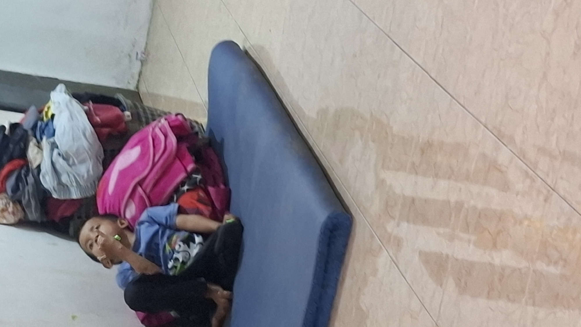 A young boy wearing a blue t-shirt and black pants is sitting on a blue mat in what appears to be a home in Cibinong Hilir, Indonesia. He is leaning against a pile of colorful laundry that includes pink and red fabrics, as well as various other clothing items. The boy has his right hand near his mouth, with a green object, possibly a toy or food item, visible. His left leg is bent and resting on the mat. The floor is tiled in a beige, marble-like pattern. The wall behind the laundry is white and appears to have some smudges. The lighting suggests it is indoors, possibly during the day or with artificial light.